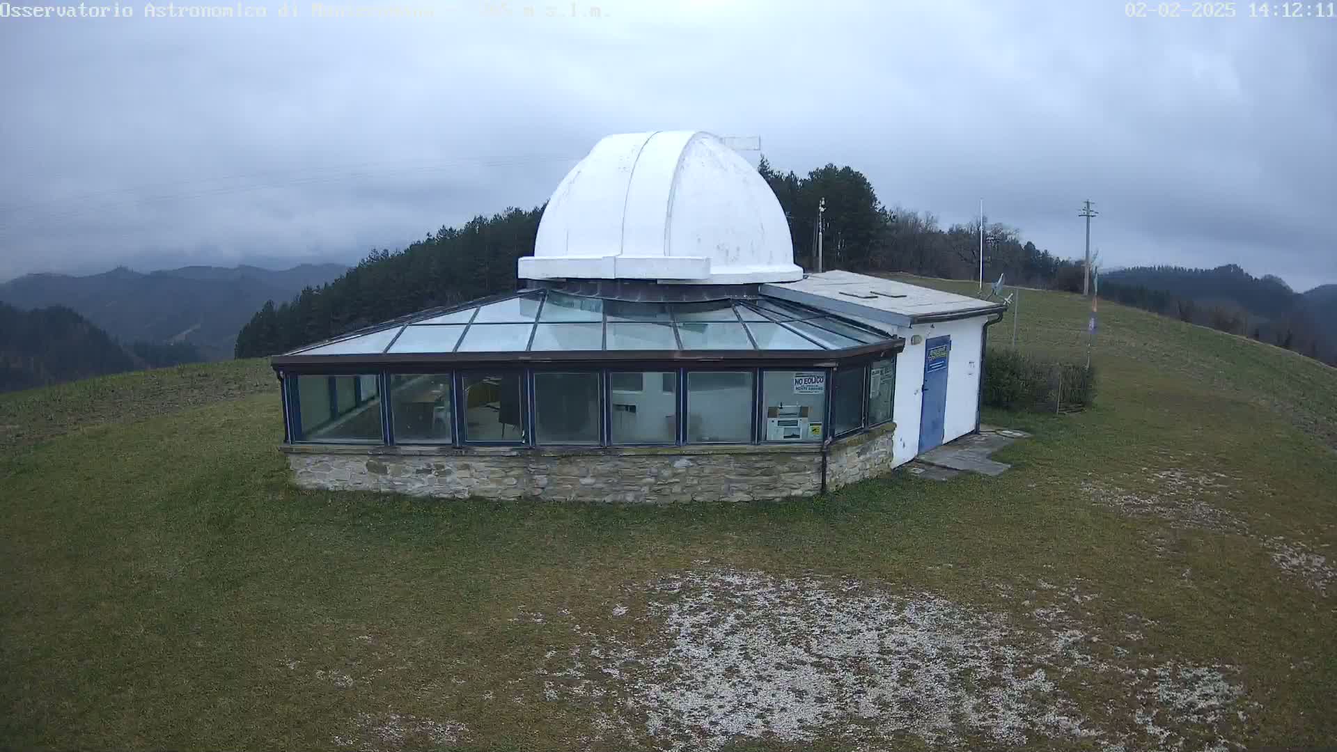 A white observatory dome sits atop a stone building with a glass sunroom, overlooking a grassy hill and distant mountains under a cloudy sky.