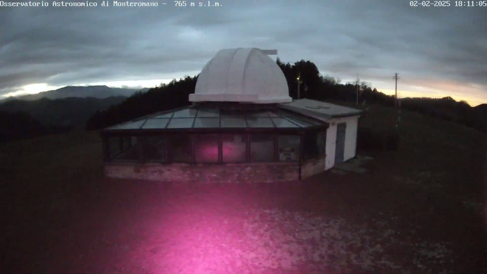 A white observatory dome sits atop a building with a glass sunroom, overlooking a dark, hilly landscape under a cloudy twilight sky.