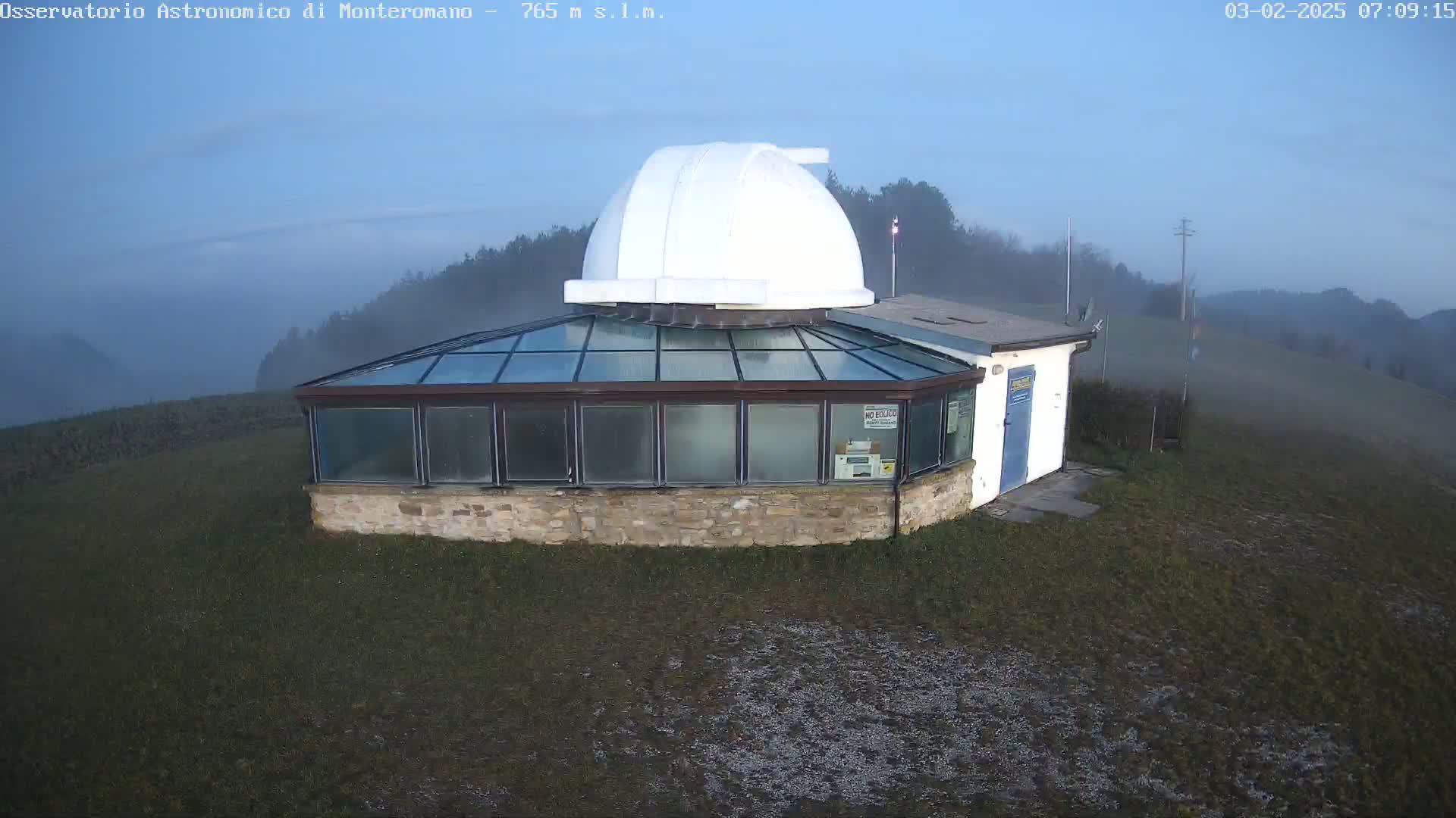 A stone observatory with a white dome sits on a grassy hill shrouded in fog under a twilight sky.