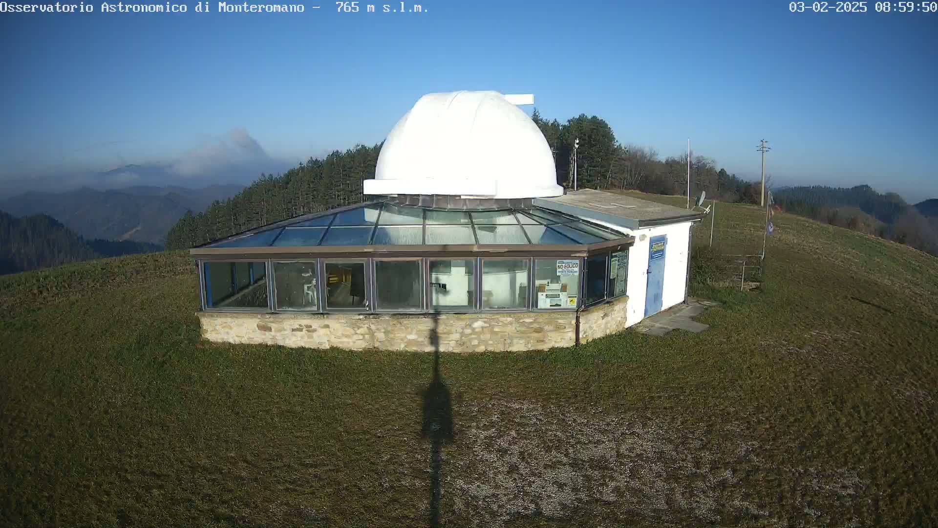 A white-domed observatory building with a glass-roofed structure sits atop a grassy hill on a clear, sunny day, with distant mountains visible in the background.