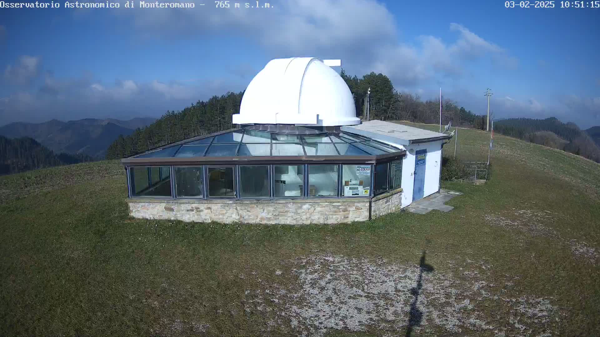 A stone observatory building with a glass enclosure and a white dome sits on a grassy hilltop under a mostly sunny sky with some scattered clouds, overlooking a mountain range in the distance.