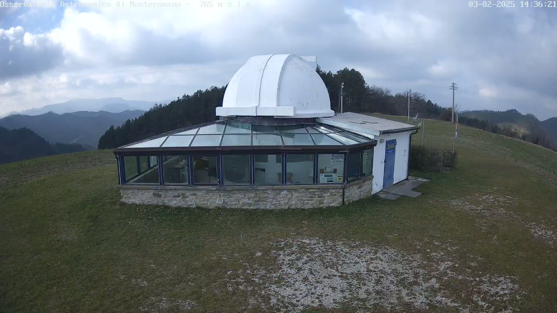 A stone building with a glass enclosure and a white dome sits on a grassy hill overlooking a mountain range under a partly cloudy sky.