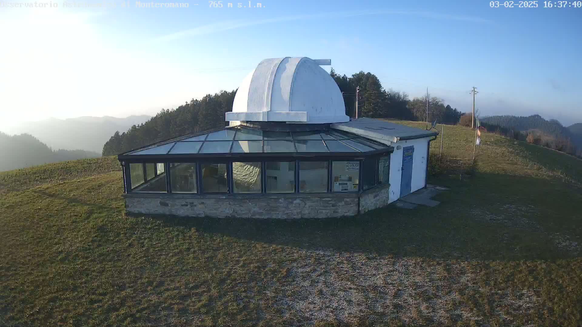 A white observatory dome sits atop a stone building with a glass enclosure, nestled on a grassy hill under a clear, sunny sky.
