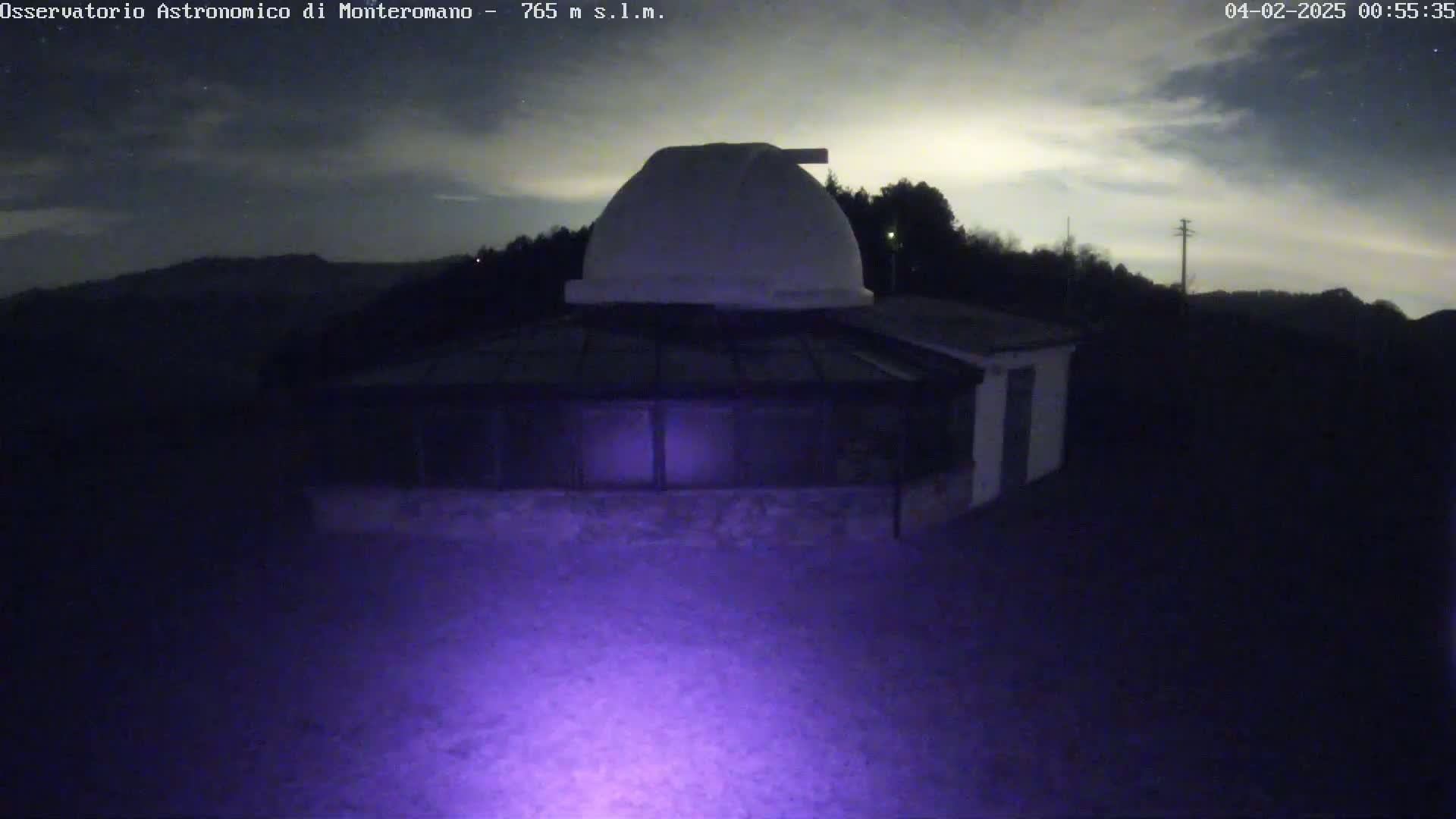 A white dome observatory building is illuminated by a purple light at night under a partly cloudy sky.