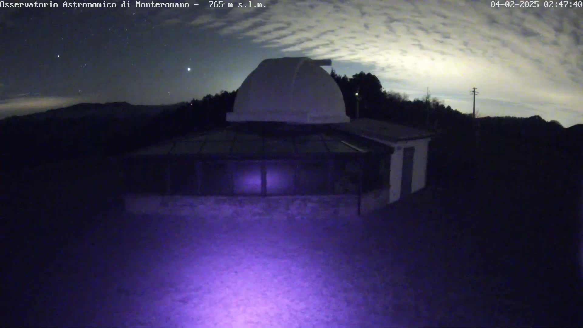 A nighttime view of a white observatory building under a partly cloudy sky, illuminated by a purple light source in the foreground.