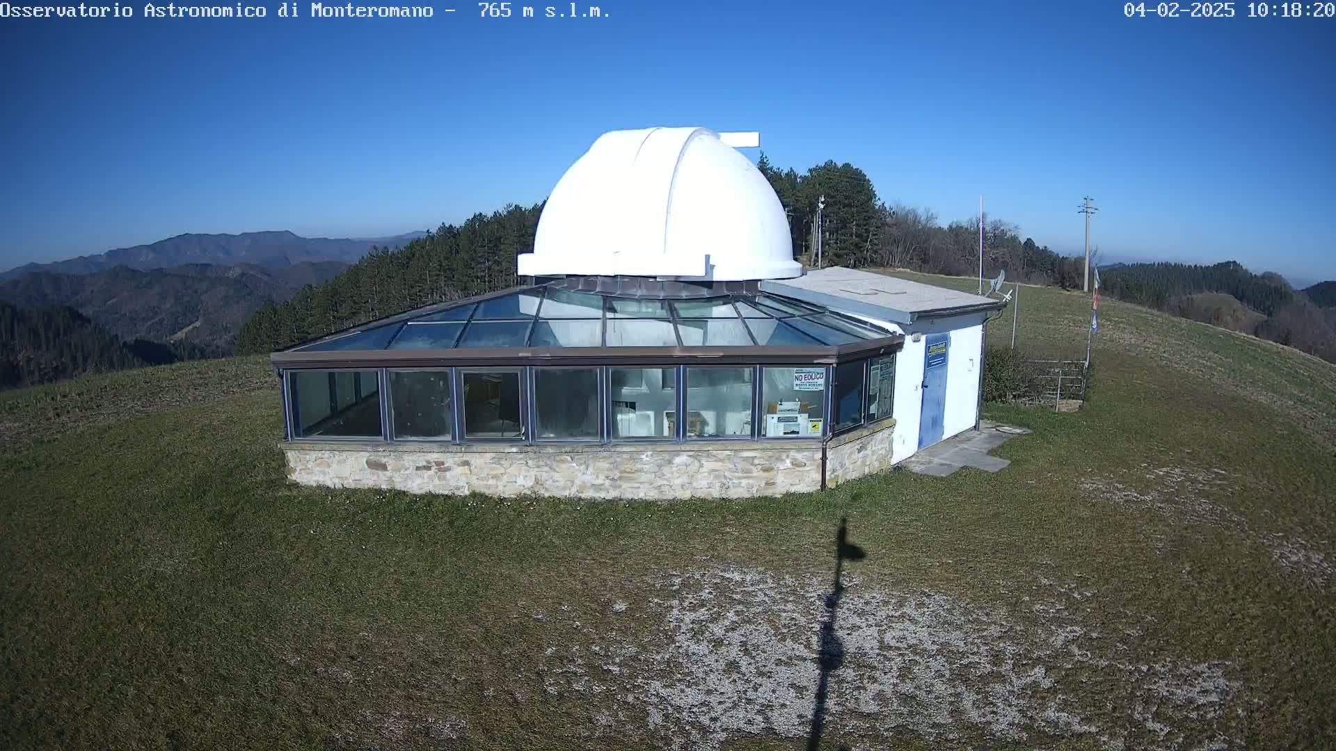 A white domed observatory building with a glass-roofed addition sits on a grassy hilltop under a clear blue sky.