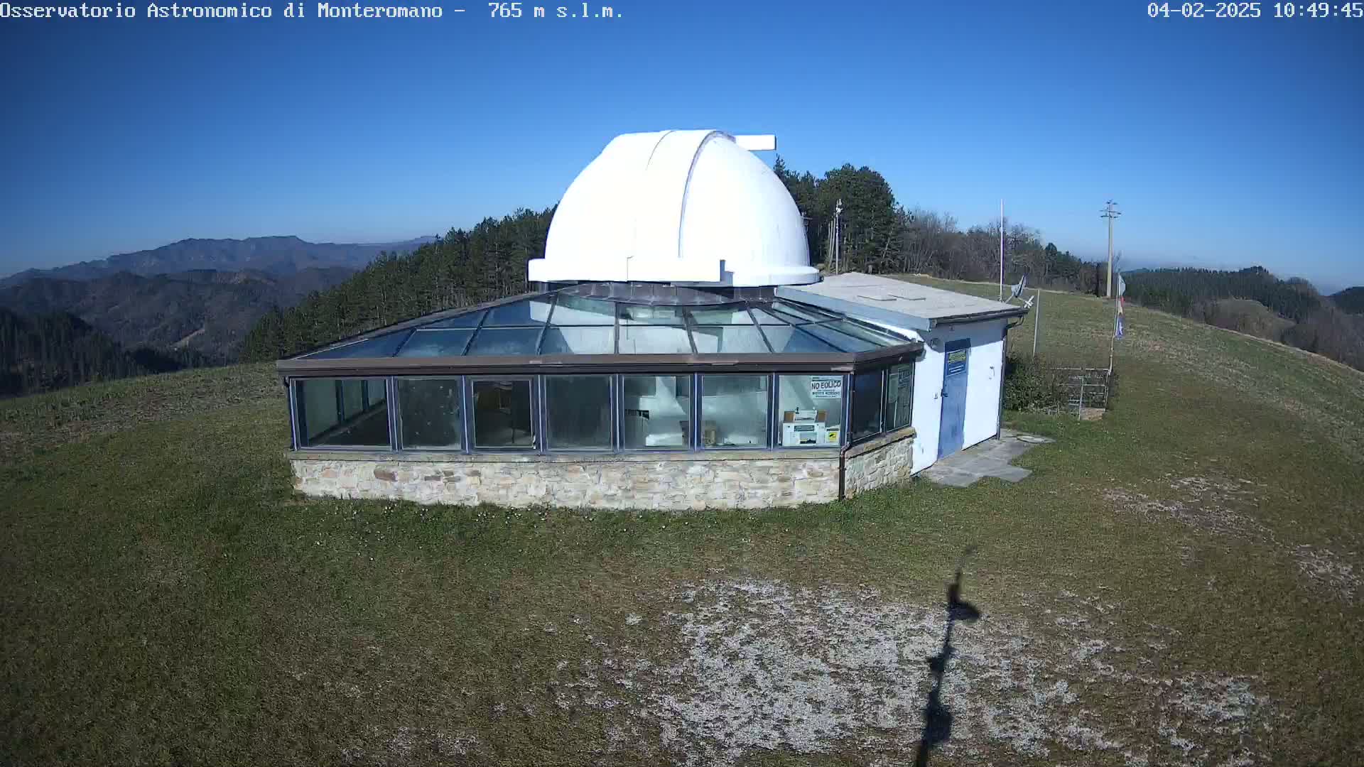A white observatory dome sits atop a stone building with a glass-roofed addition, all situated on a grassy hill under a clear blue sky.