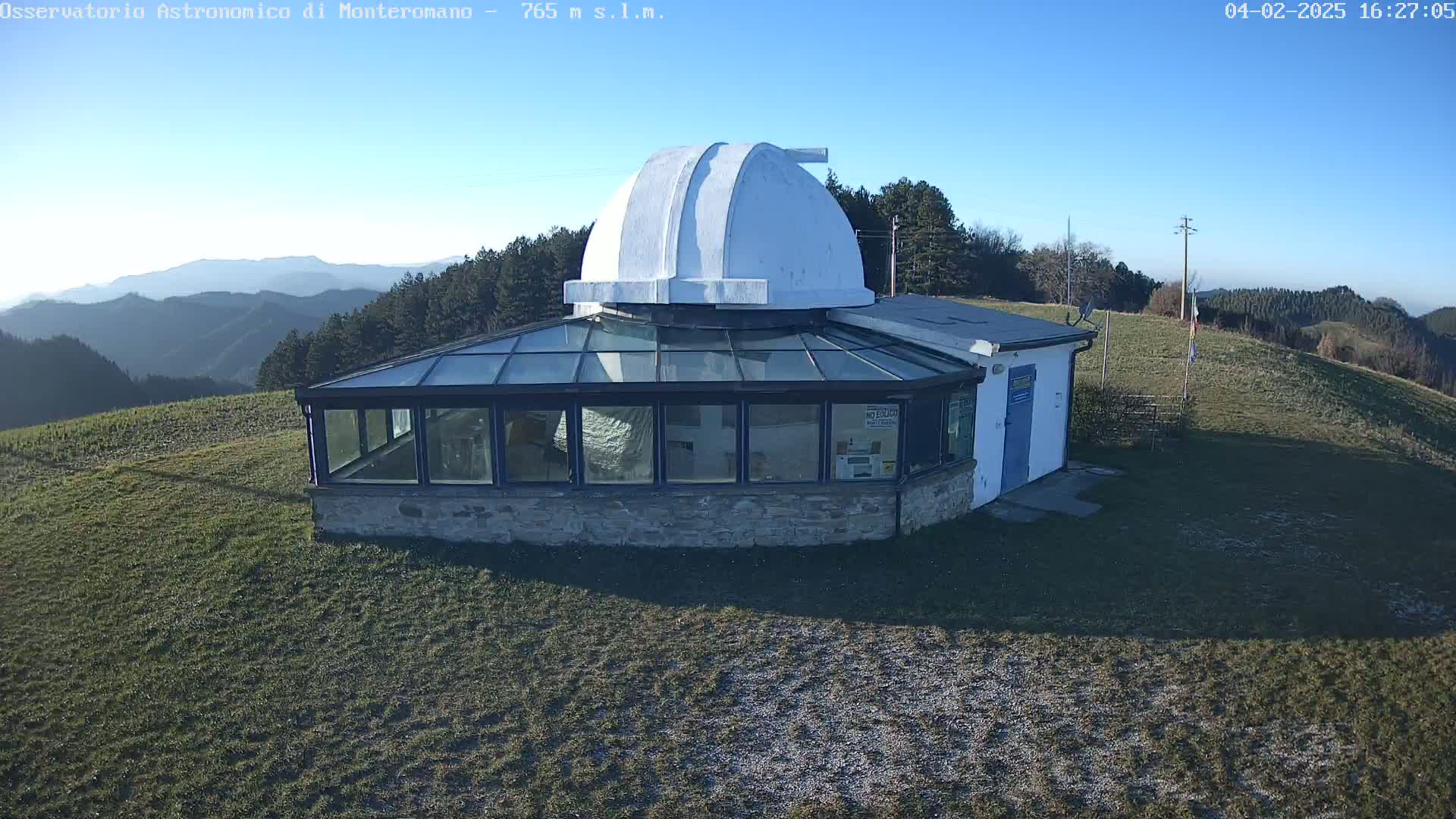 A white domed observatory building with a glass enclosure sits on a grassy hill overlooking a mountain range under a clear blue sky.