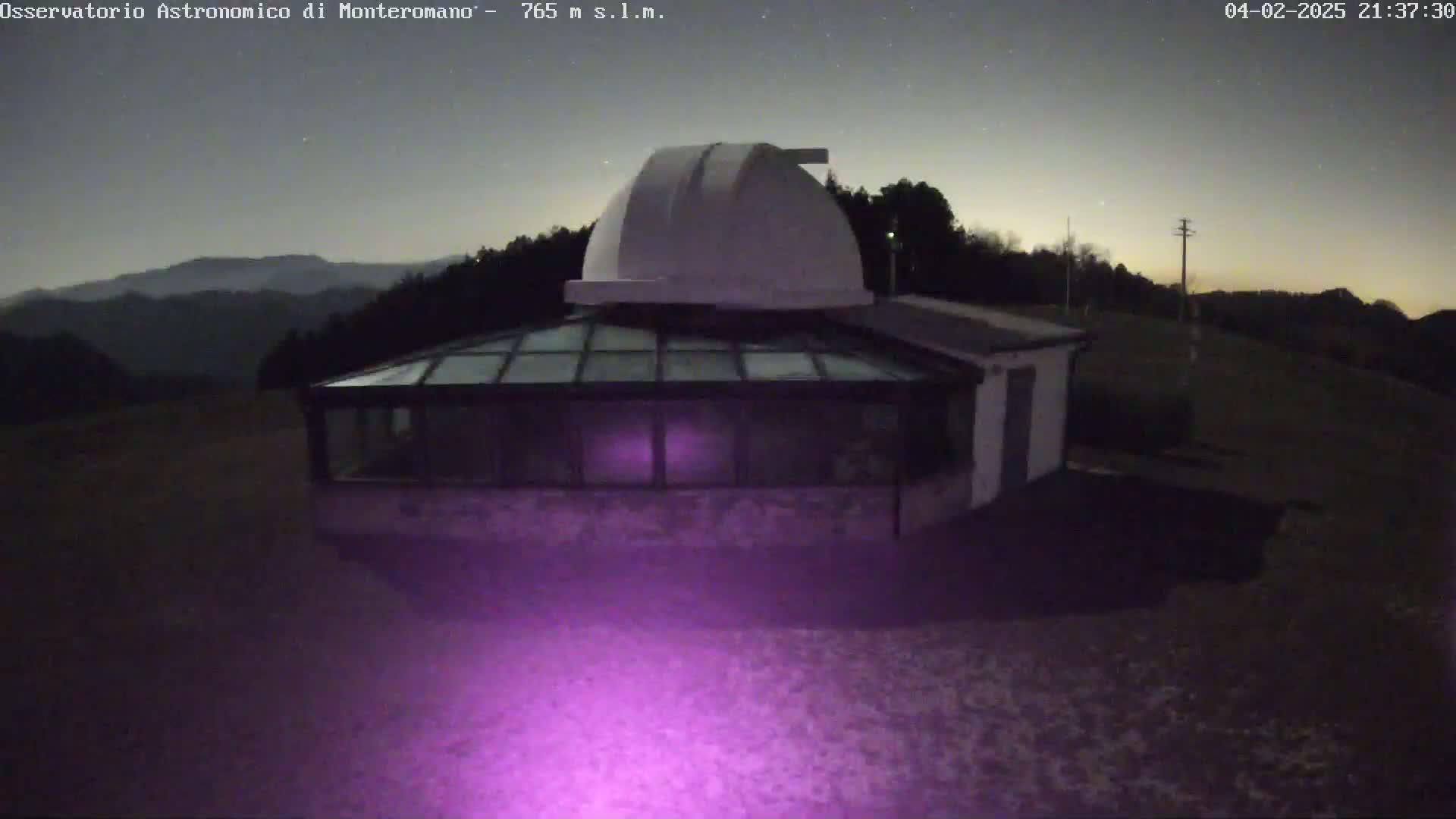 Under a clear, starry night sky, a white observatory dome sits atop a stone building with a glass-roofed addition, all situated on a hill overlooking a dark, mountainous landscape.