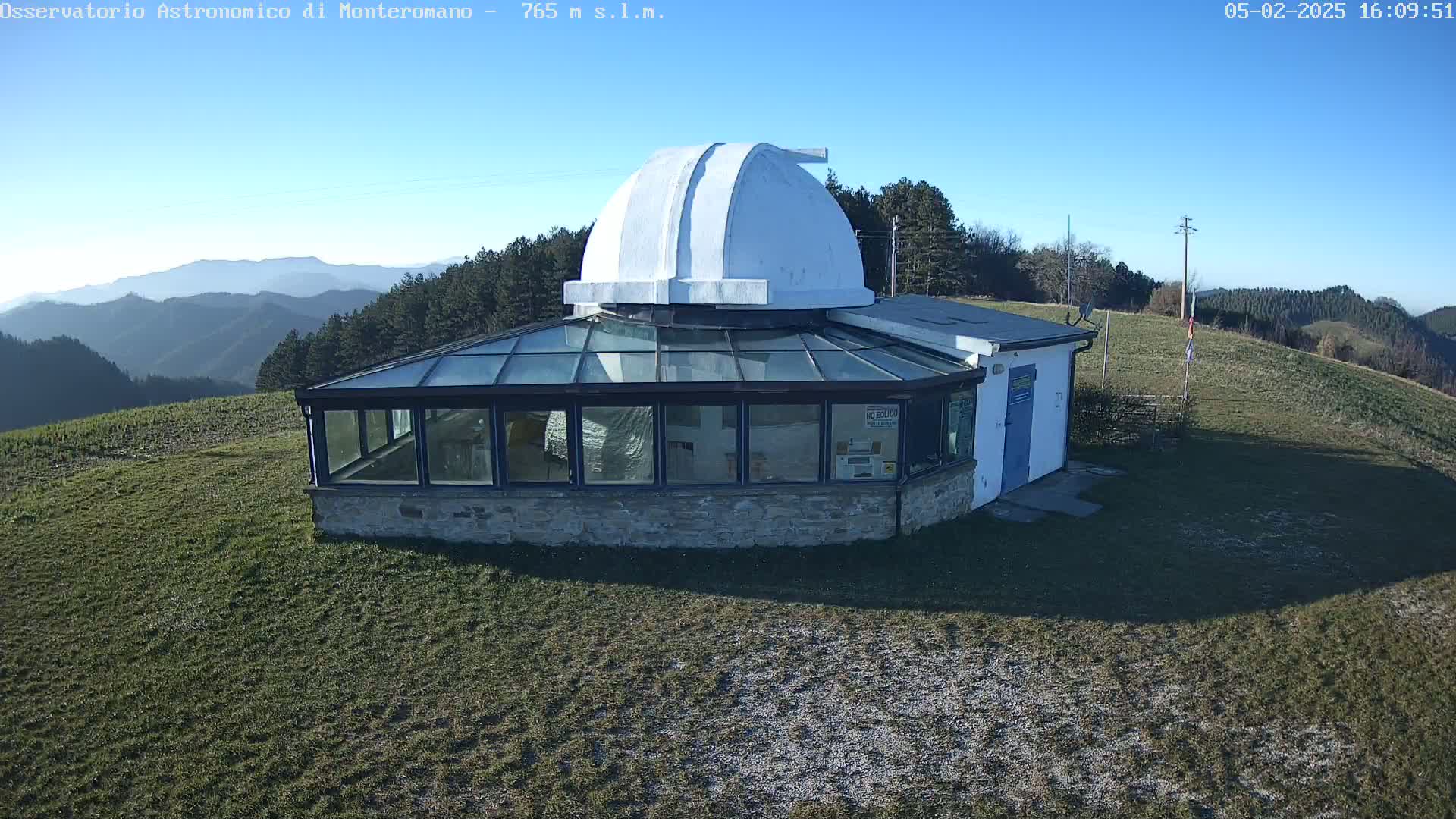 A stone and glass observatory building with a white dome sits atop a grassy hill on a clear, sunny day, overlooking a mountain range in the distance.