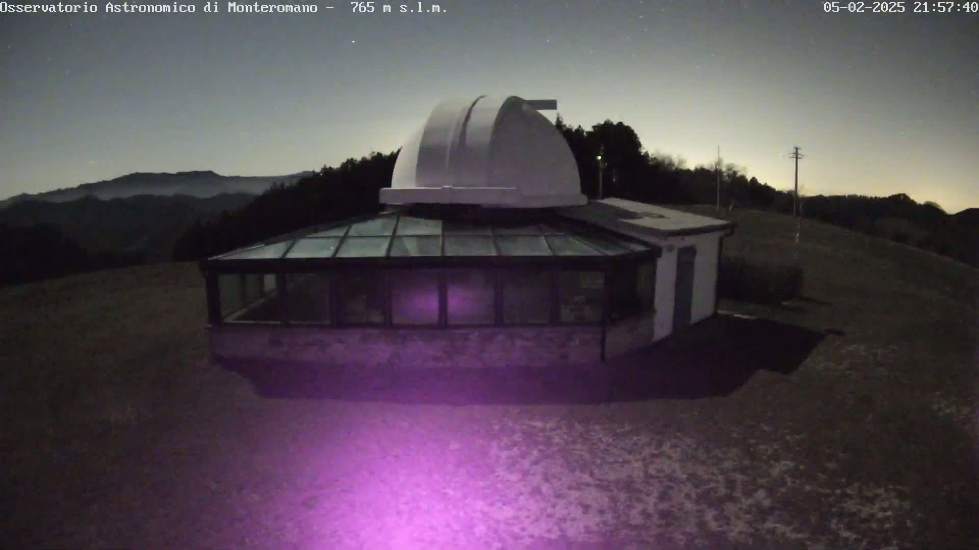 A white domed observatory building with a glass-roofed addition sits on a dark, lightly snow-covered hill under a night sky.