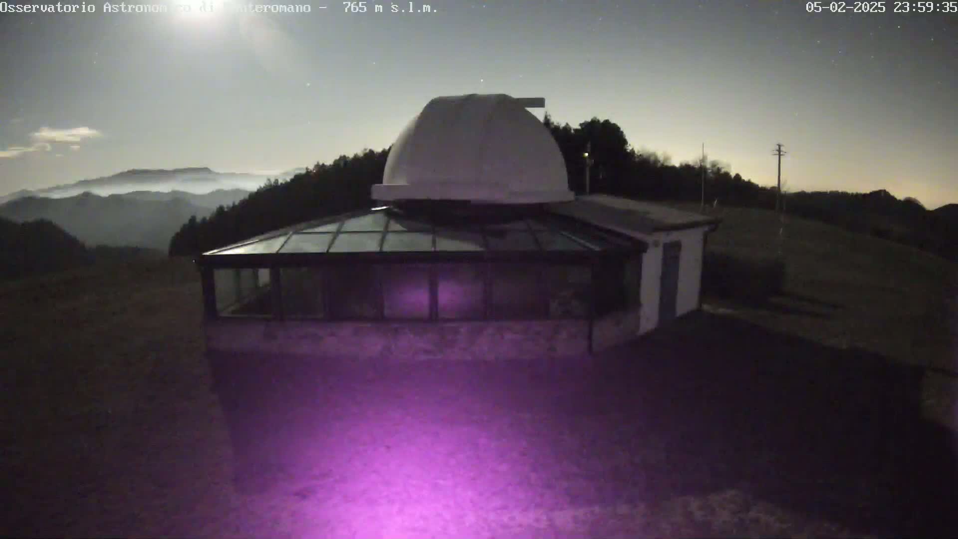 A nighttime view of an observatory building with a dome, situated on a hill overlooking a hazy valley under a clear, starry sky.
