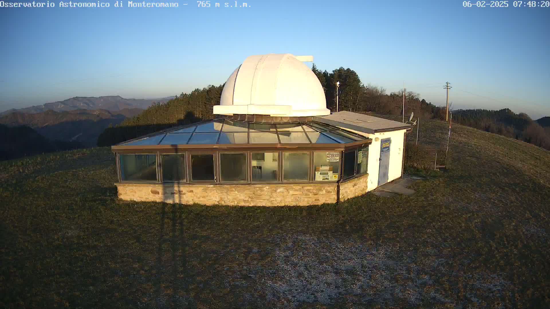 A stone building with a glass roof and a large white dome sits atop a grassy hill under a clear blue sky.