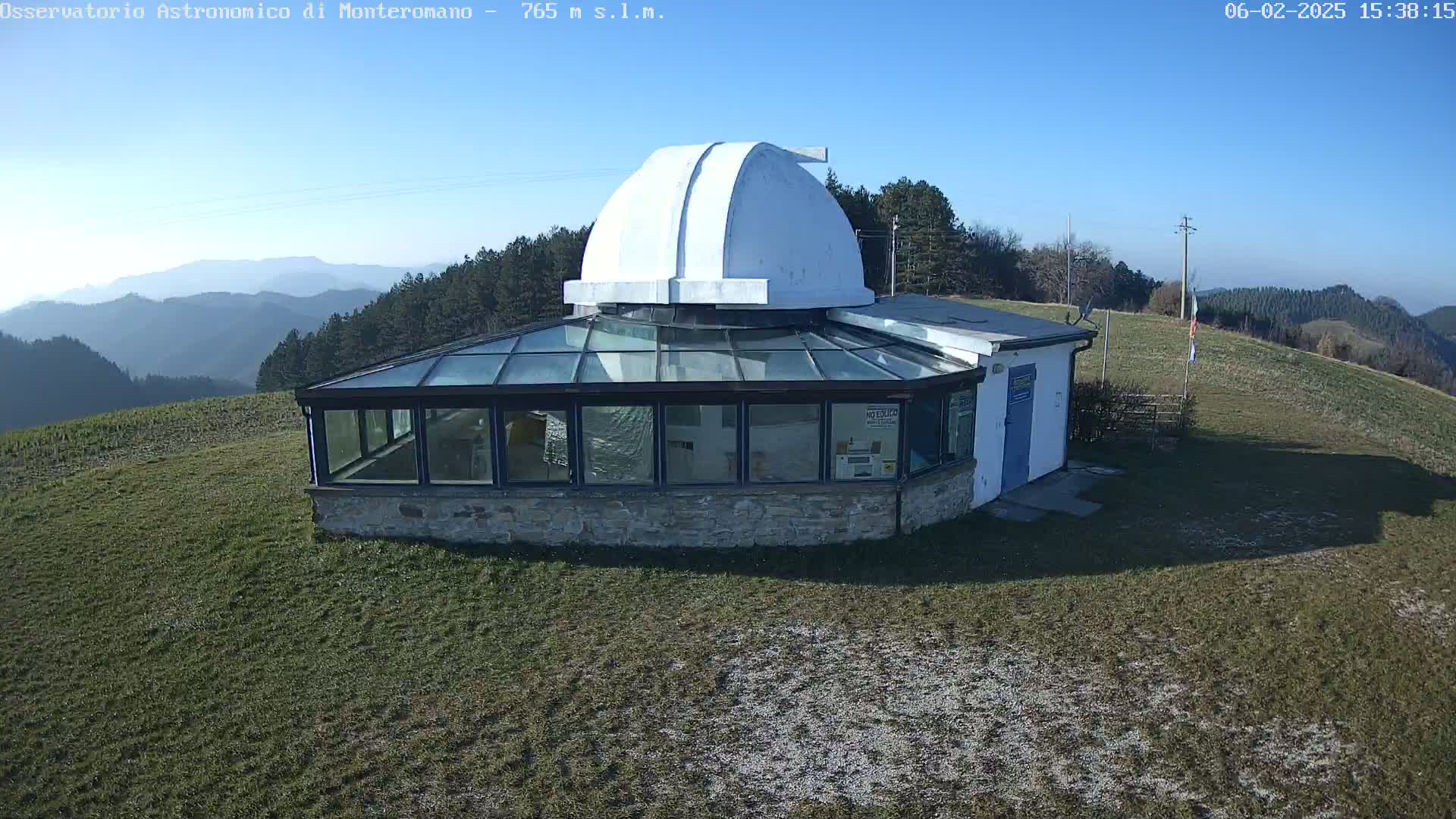 A stone building with a glass enclosure and a white dome sits atop a grassy hill on a clear, sunny day, overlooking a range of distant hills.