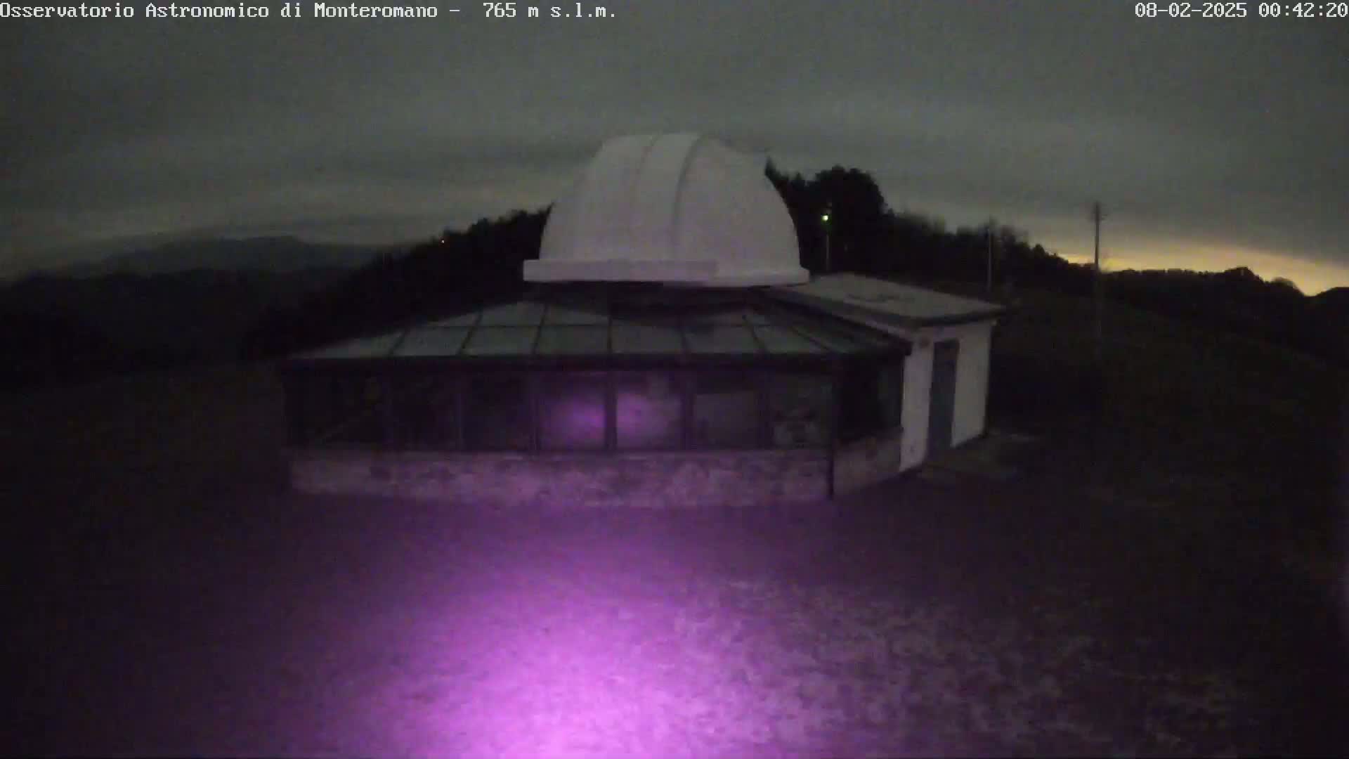 A white domed observatory building with a glass-roofed addition is illuminated by a purple light at night under a dark, cloudy sky.