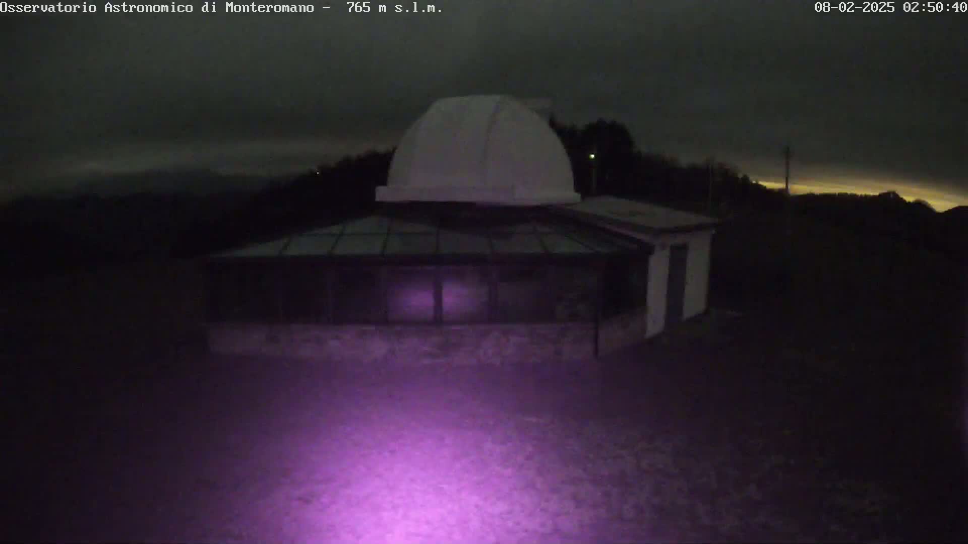 A low-light nighttime image shows an observatory building with a dome on top, under a dark, cloudy sky with a hint of light on the horizon.