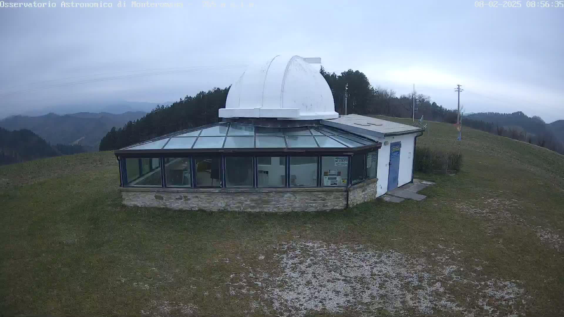A stone building with a glass conservatory and a white dome on top sits on a grassy hill overlooking a hazy mountain range under an overcast sky.