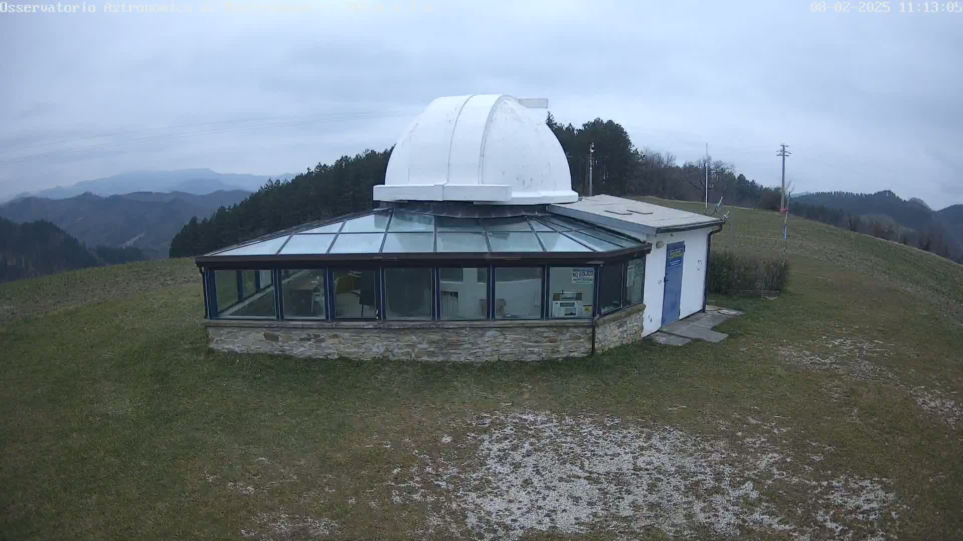 A stone observatory with a glass enclosure and a white dome sits on a grassy hill overlooking a range of mountains under a cloudy sky.