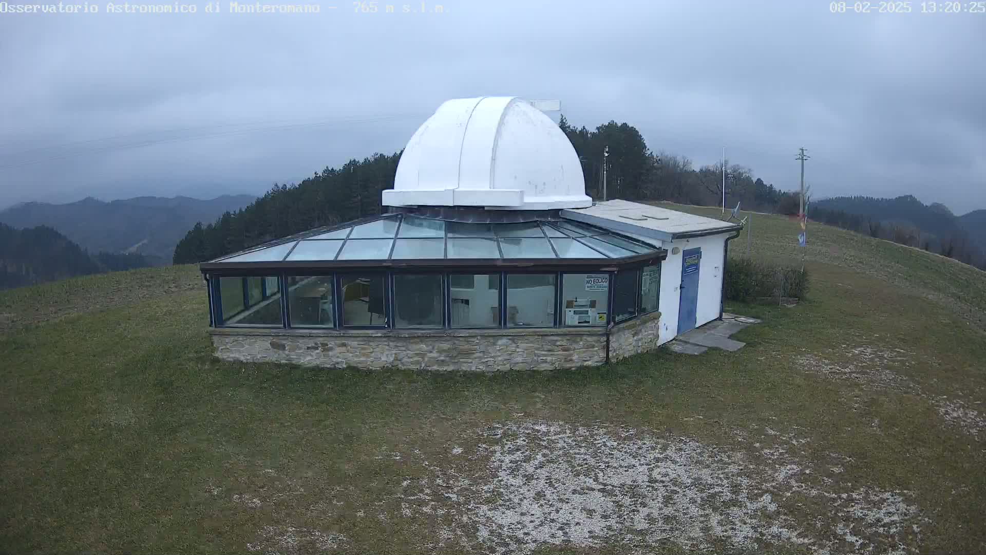 A stone building with a glass enclosure and a white dome on top sits atop a grassy hill overlooking a hazy mountain range under an overcast sky.