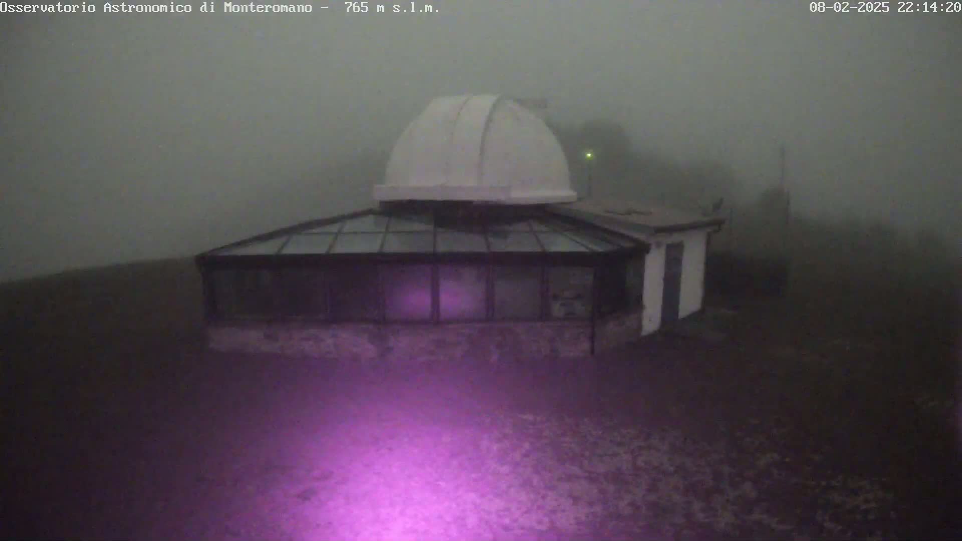 A white dome-shaped observatory building is shrouded in thick fog at night, illuminated by a purplish light emanating from the ground.