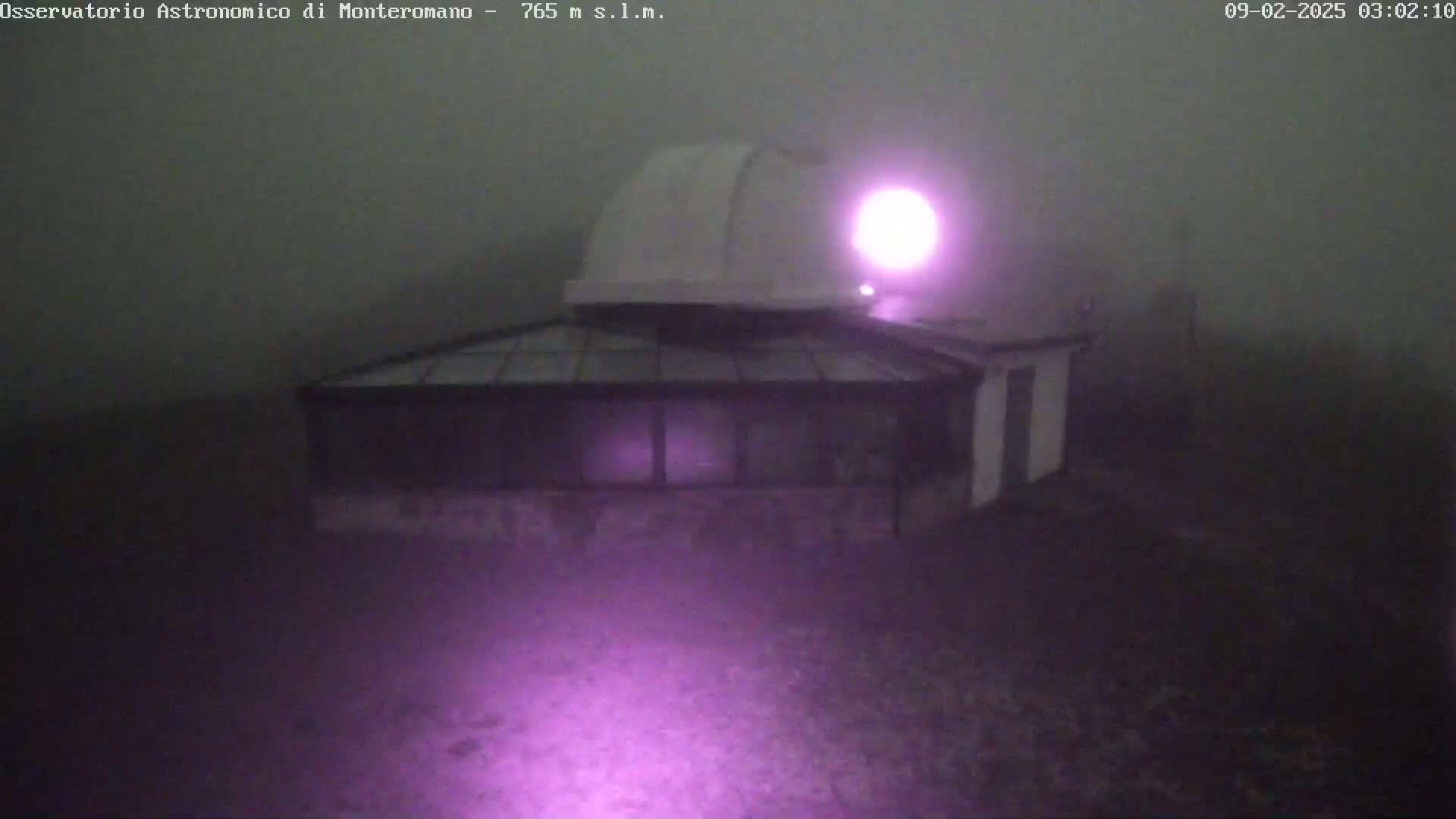 A white observatory dome and attached building are shrouded in heavy fog at night, illuminated by a purple light.