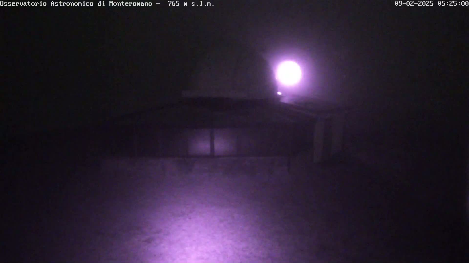 A low-light image shows a building at night in foggy conditions, illuminated by a bright light source.