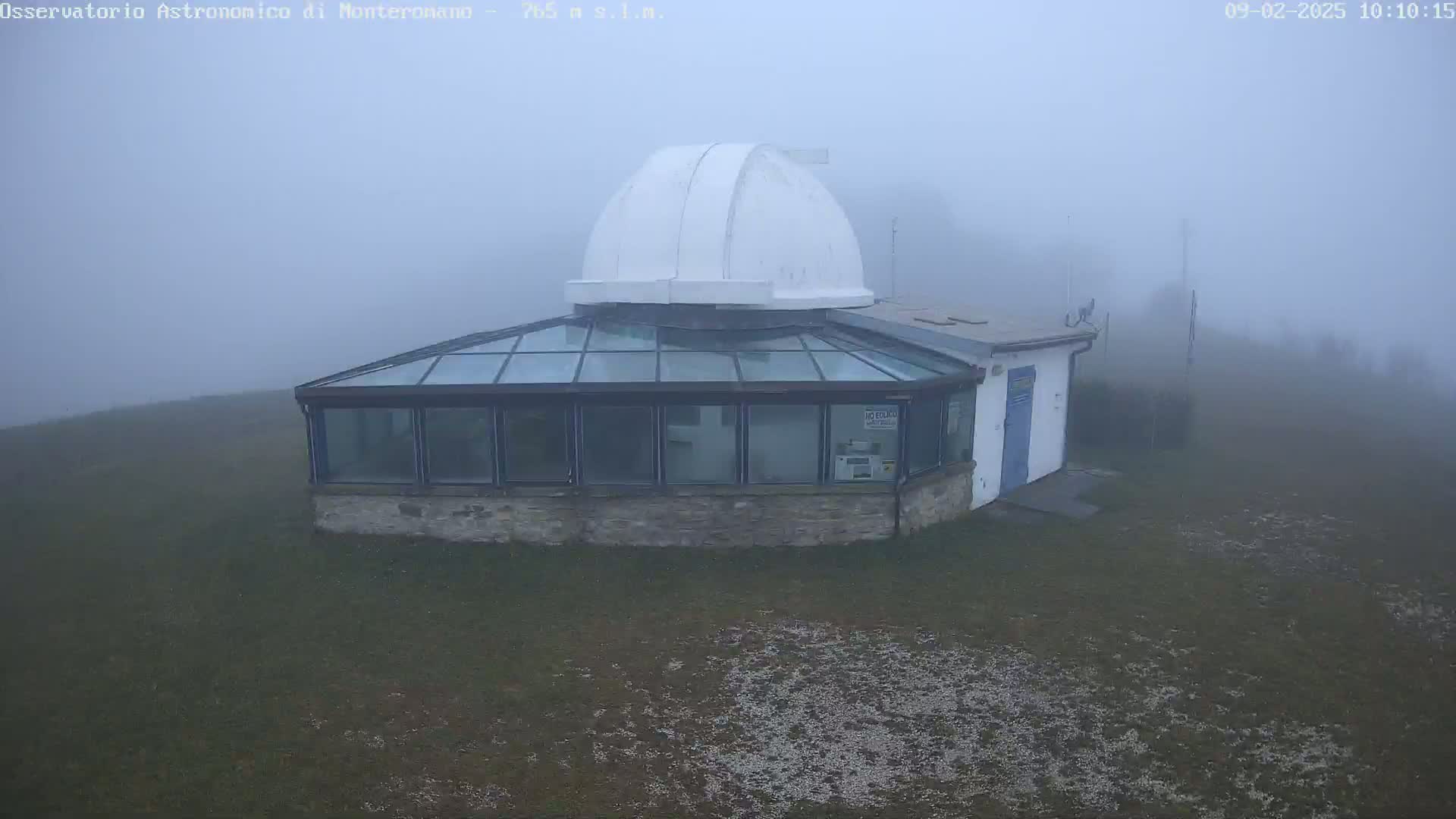 A white-domed observatory building with a glass-roofed addition sits on a grassy hill shrouded in thick fog.