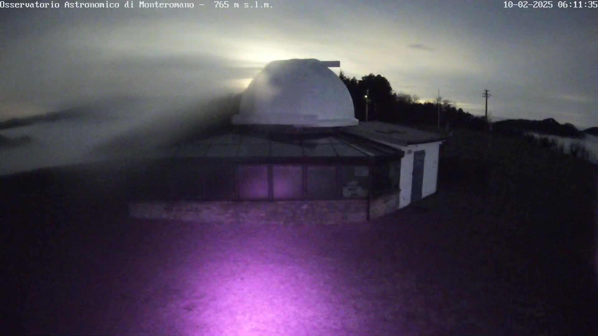 A white observatory dome and attached building are visible under a twilight sky with some fog or low clouds.