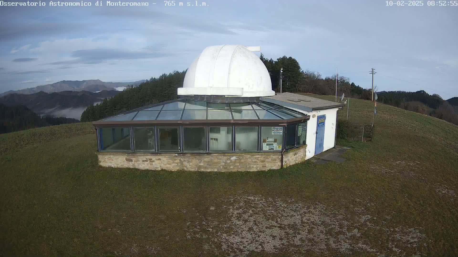 An observatory with a white dome sits atop a grassy hill on a partly cloudy day, overlooking a valley partially shrouded in fog.