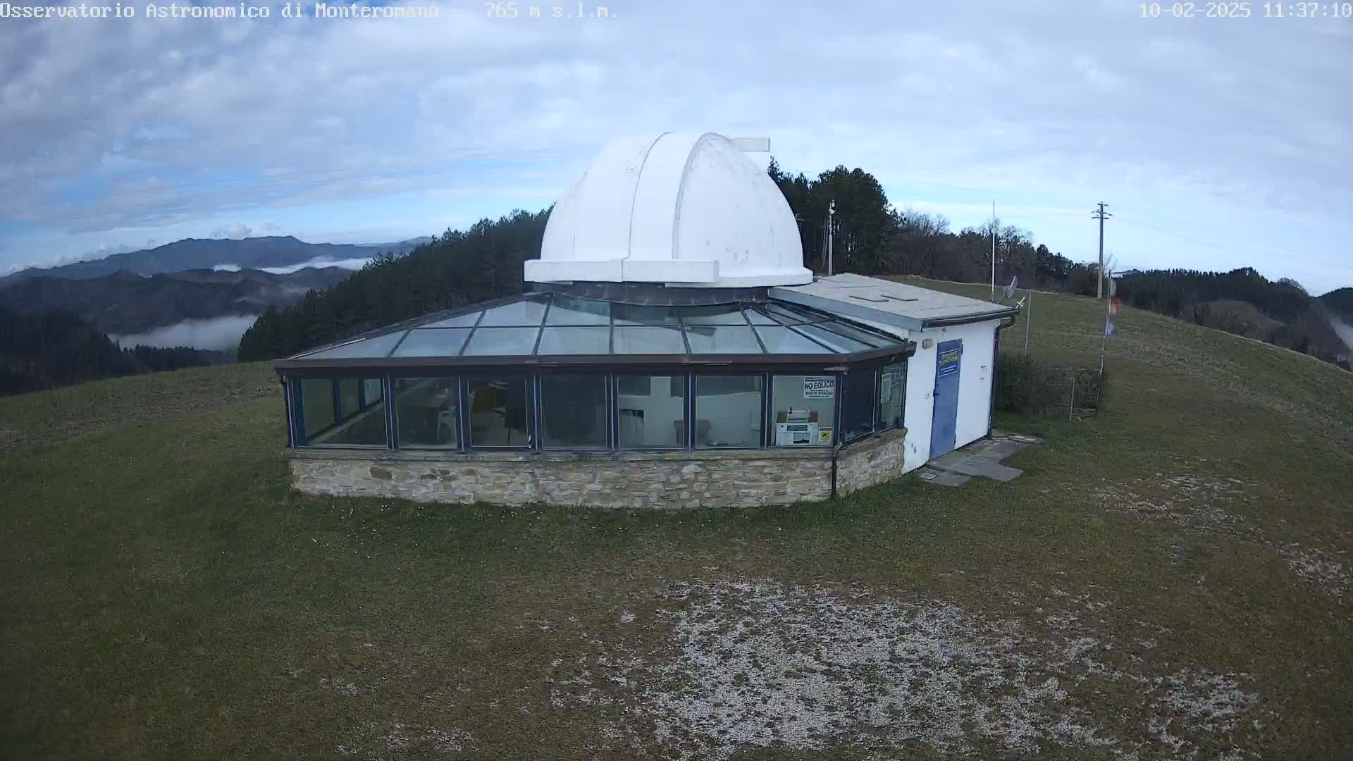 An observatory with a white dome sits on a grassy hilltop overlooking a valley partially covered in fog under a mostly cloudy sky.