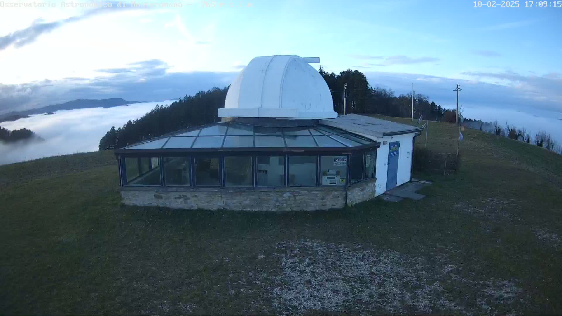 A stone building with a glass conservatory and a white observatory dome sits atop a hill overlooking a valley filled with fog under a mostly clear sky.