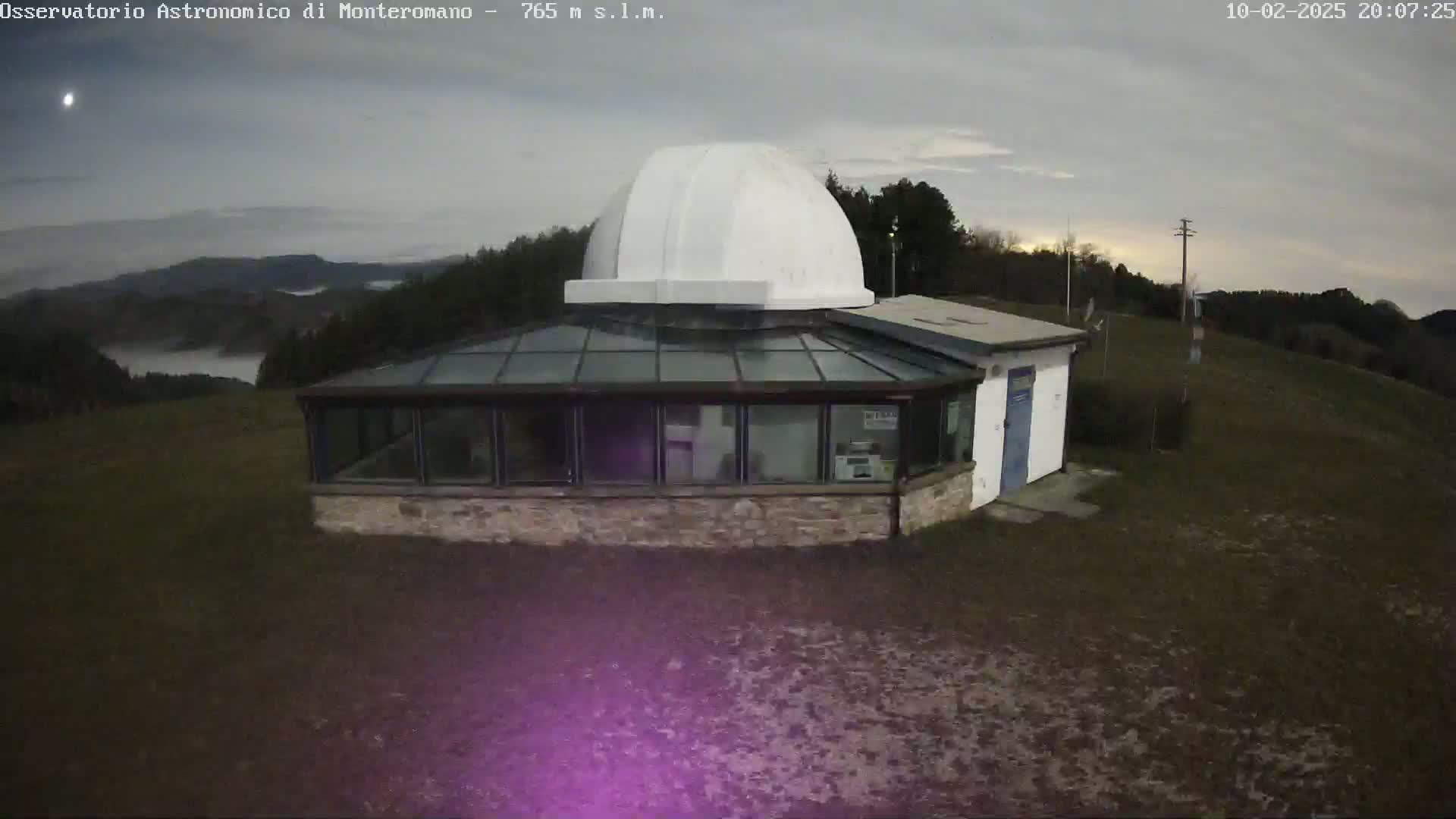 A white-domed observatory building with a glass-roofed addition sits on a hill overlooking a valley shrouded in fog under a cloudy sky.