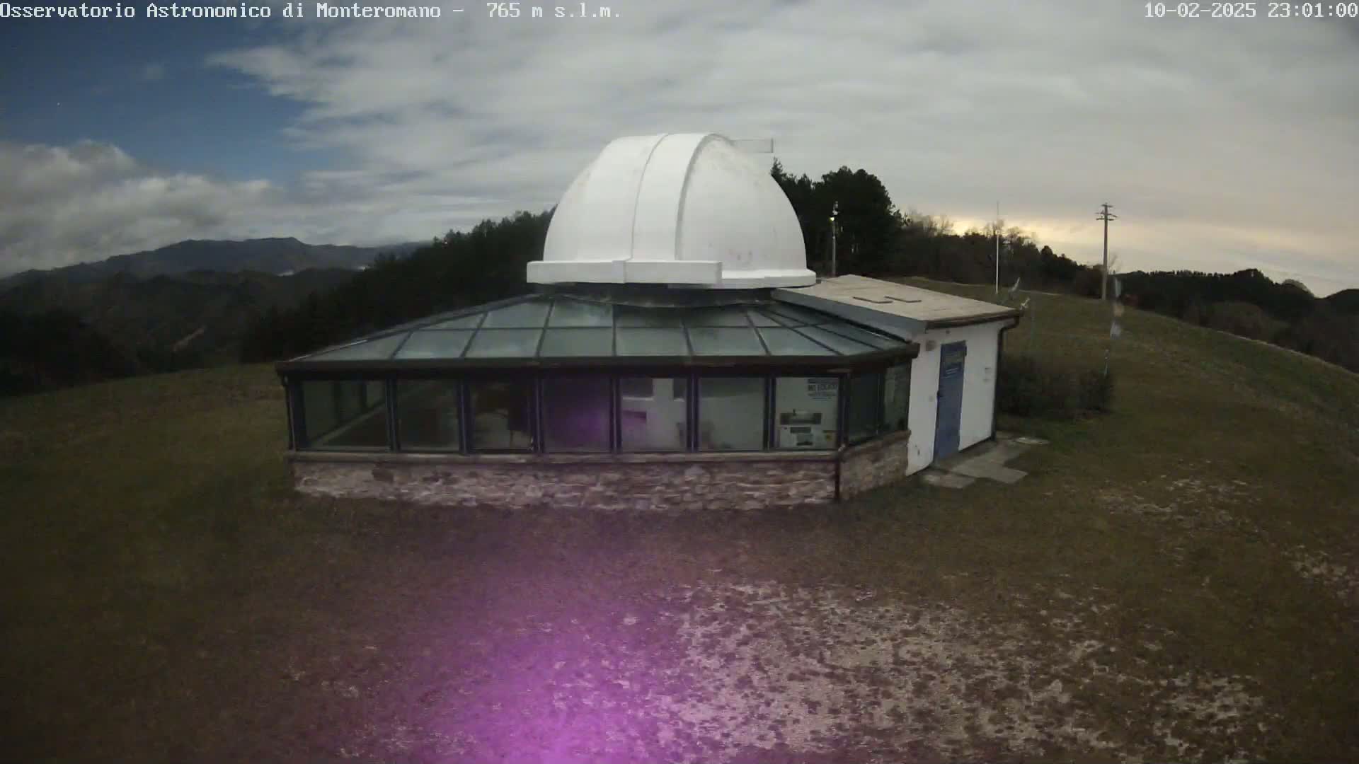 A white dome observatory building with a glass-roofed addition sits on a hilltop under a partly cloudy night sky.