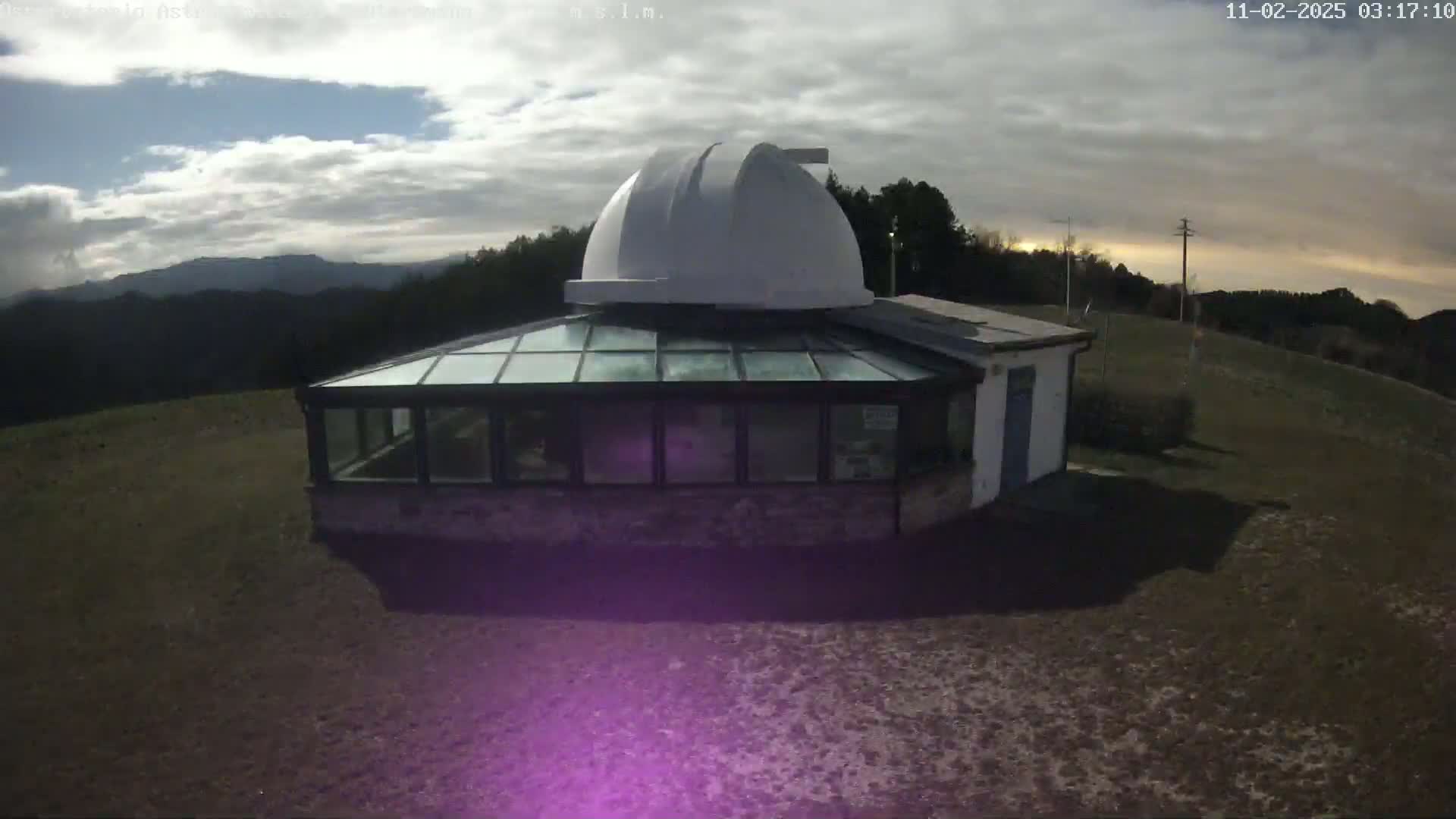 A small observatory with a glass-roofed addition sits on a grassy hilltop under a partly cloudy sky.