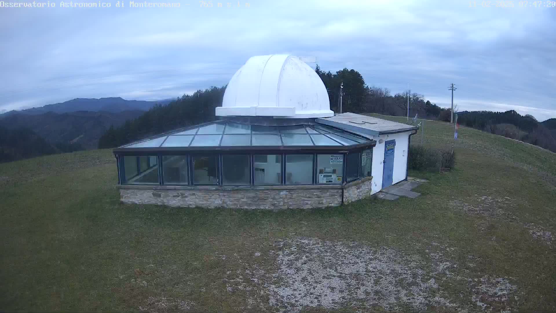 An observatory with a white dome sits atop a hill on a cloudy day, overlooking a range of mountains in the distance.