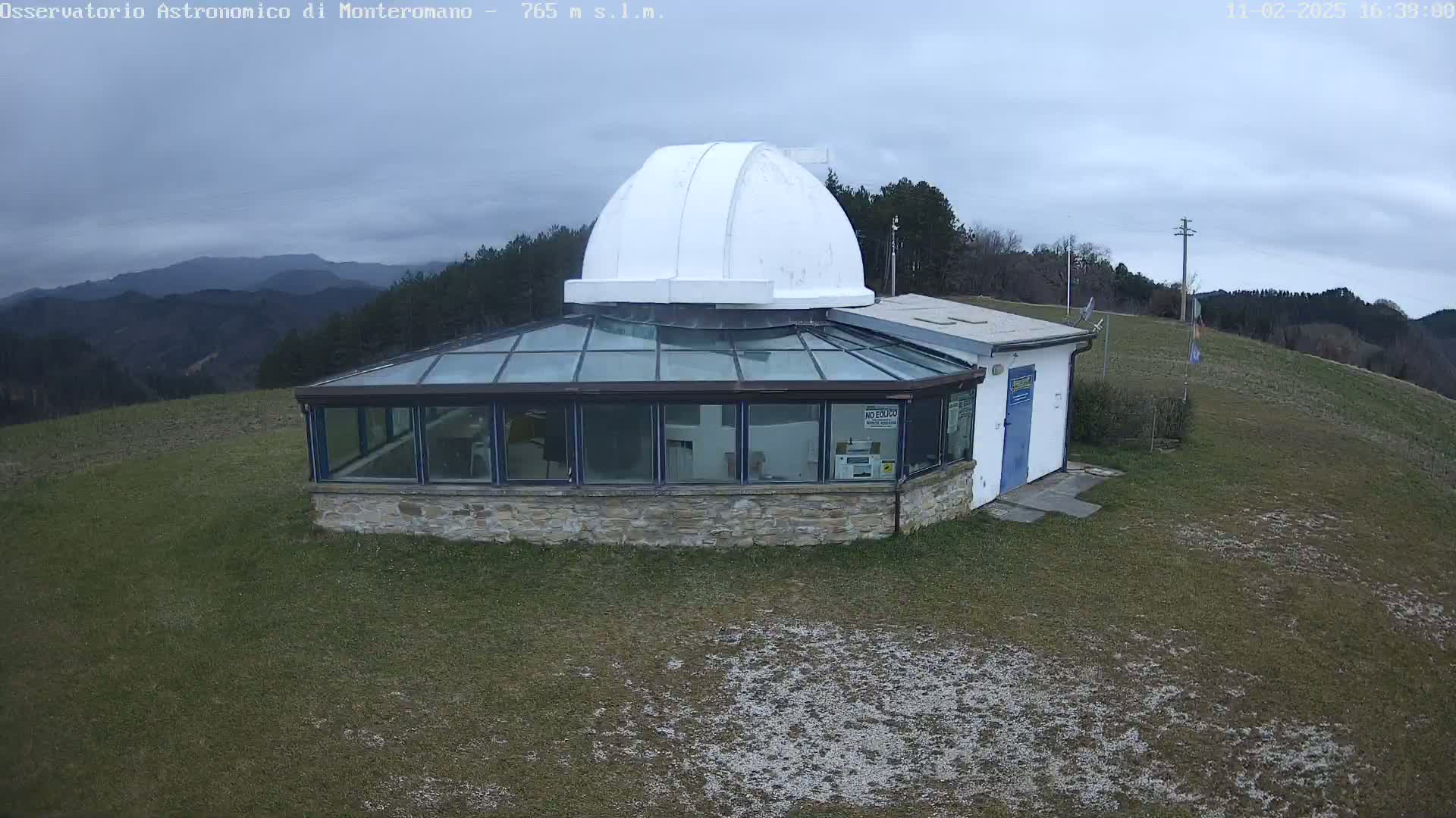 A white domed observatory building with a glass-roofed addition sits on a grassy hilltop under an overcast sky, overlooking a range of distant hills.