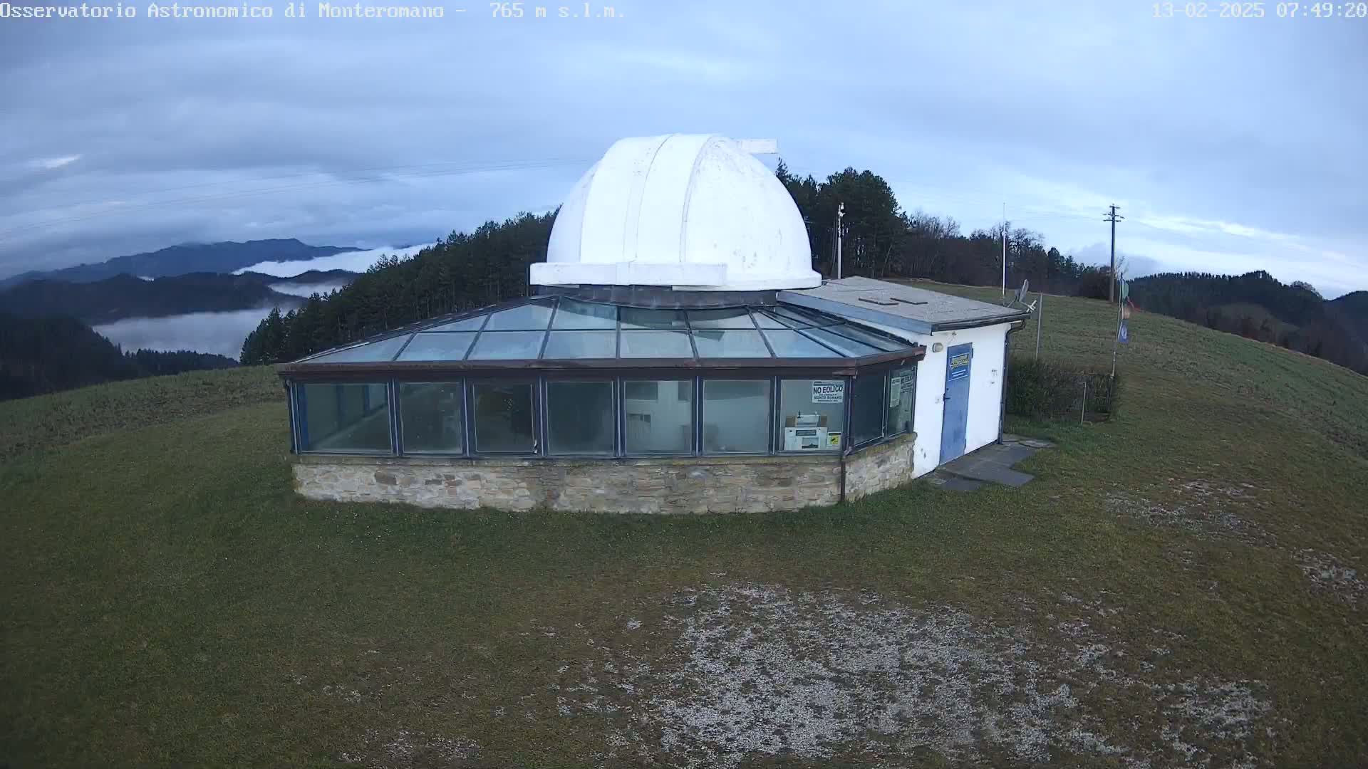 An observatory with a white dome sits on a grassy hilltop overlooking a valley filled with low-lying fog under a mostly cloudy sky.