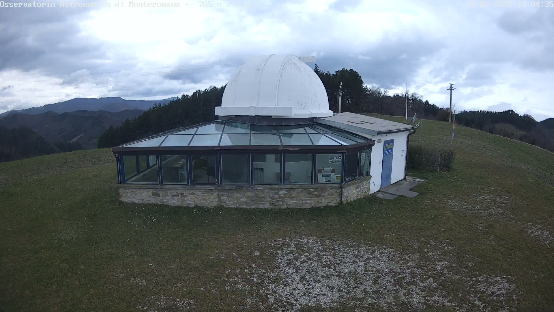 A stone building with a glass roof and a white dome on top sits on a grassy hill under a cloudy sky, overlooking a mountain range in the distance.