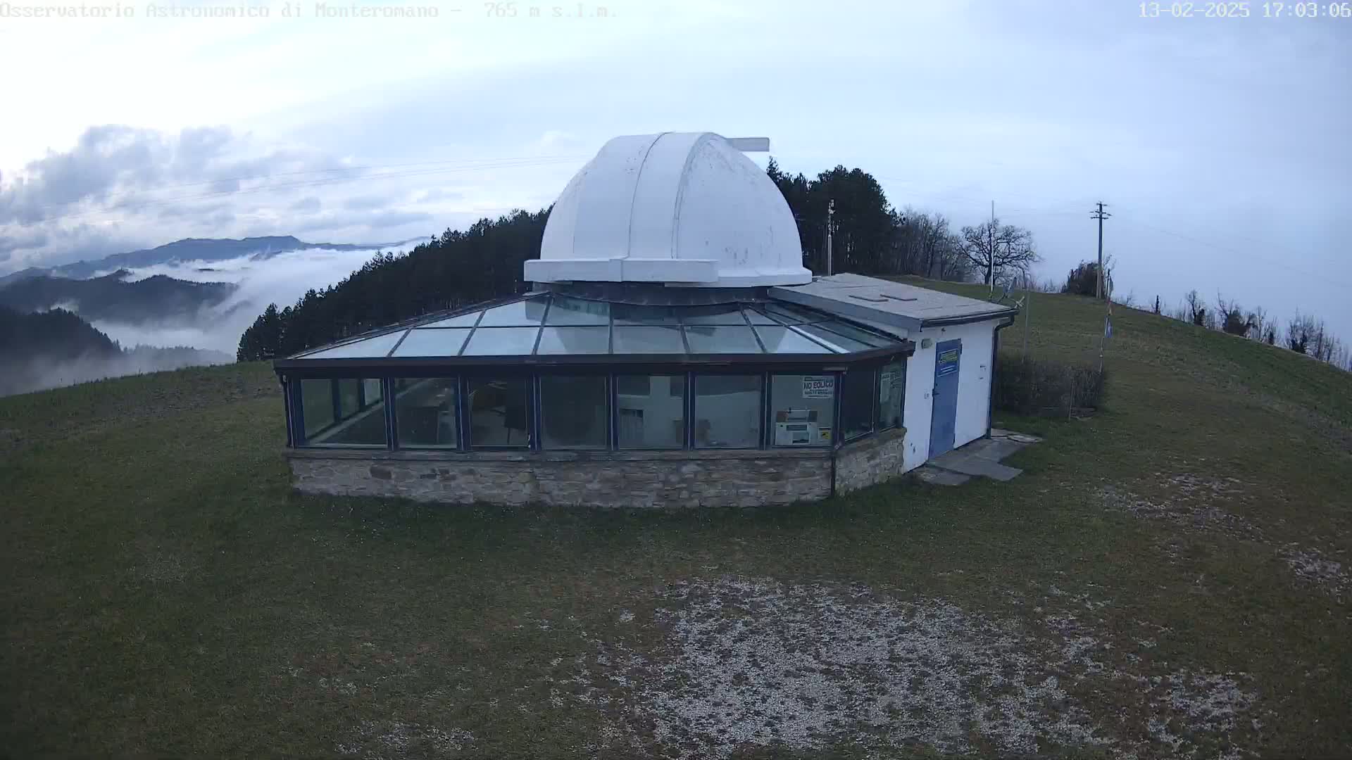 An observatory with a glass-enclosed section and a white dome sits on a grassy hill overlooking a valley shrouded in fog under a cloudy sky.