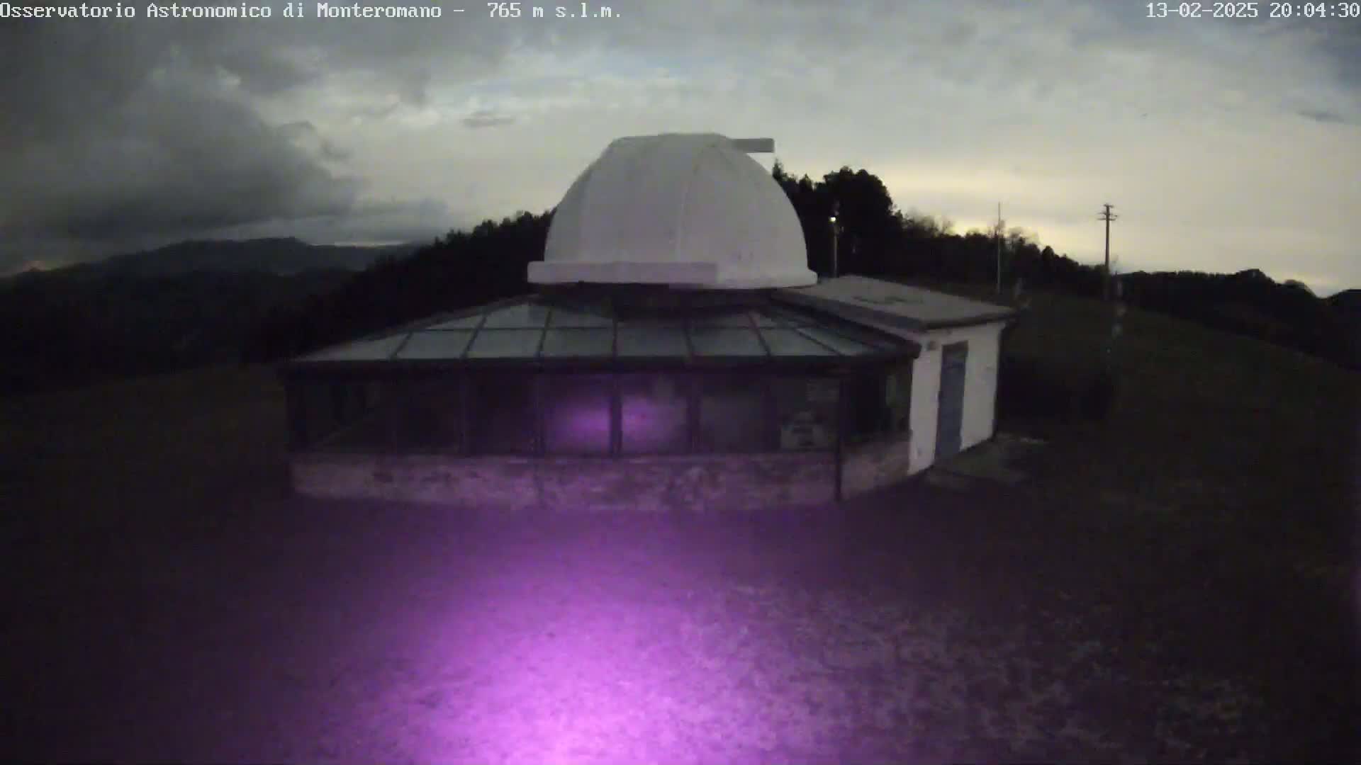 A white domed observatory building with a glass-roofed addition is situated on a hilltop under a partly cloudy, dusky sky.