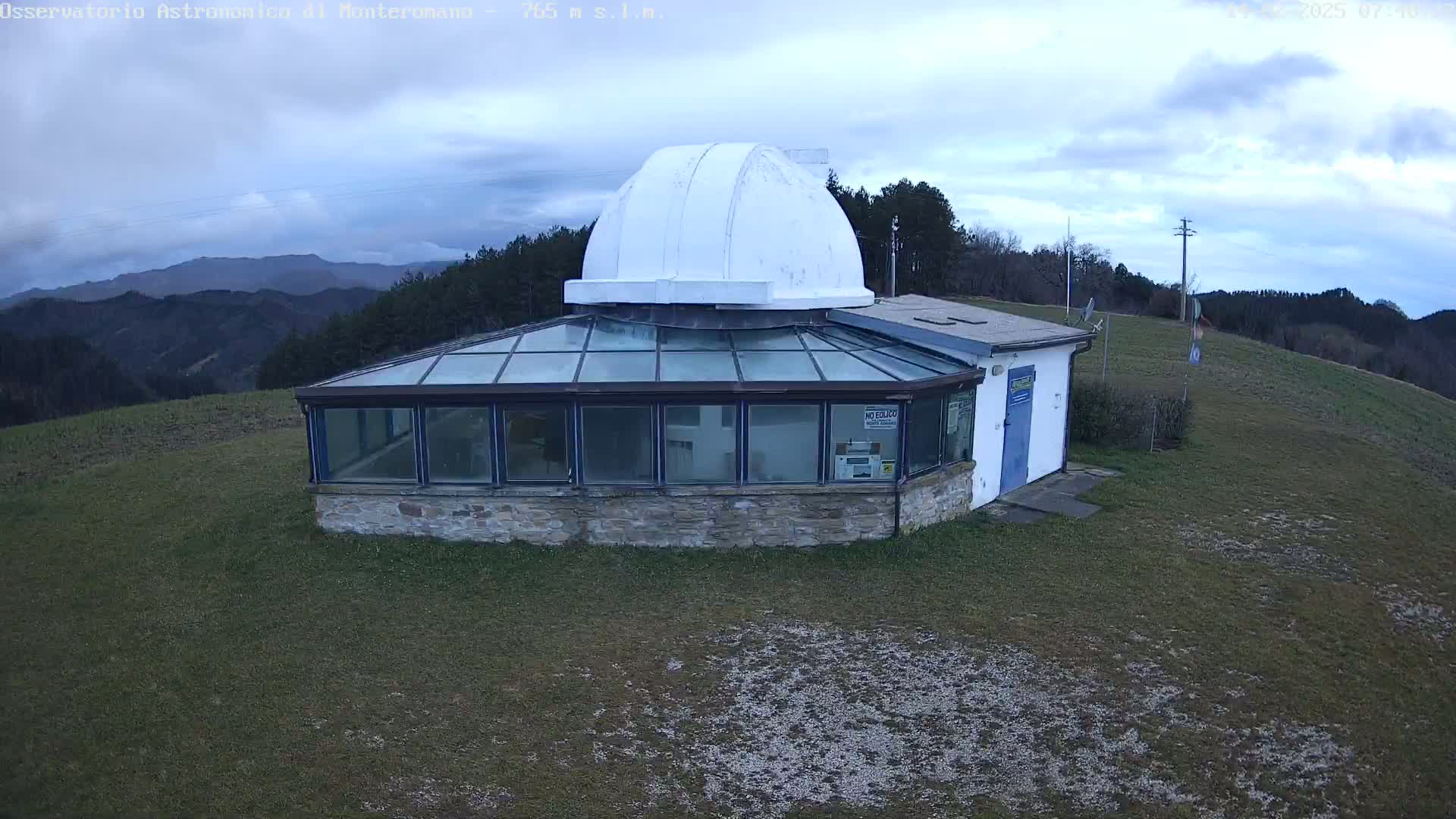 A stone building with a glass enclosure and a white dome on top sits on a grassy hill under a cloudy sky.