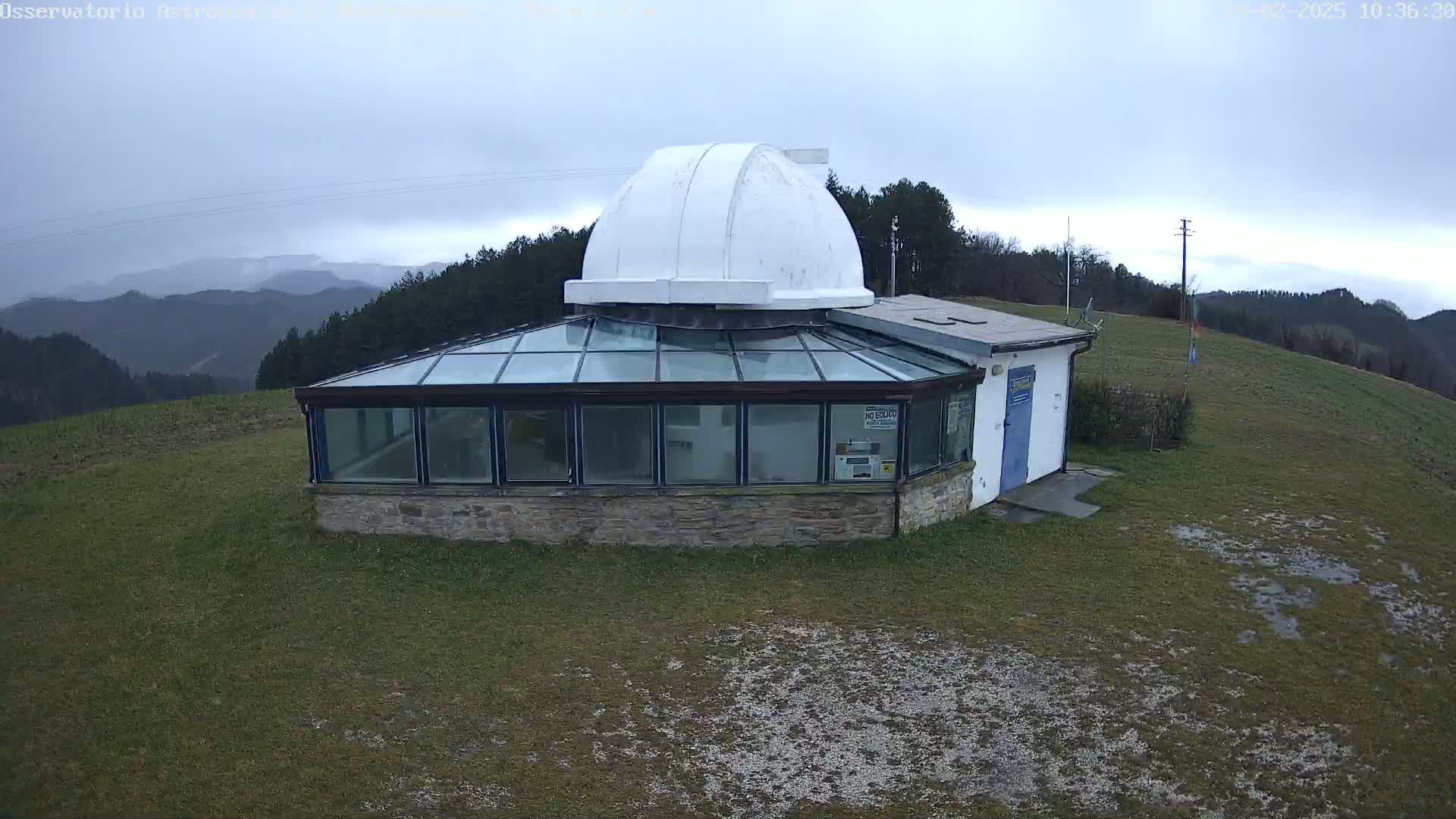 A stone building with a glass conservatory and a white dome sits on a grassy hill overlooking a range of mountains under an overcast sky.