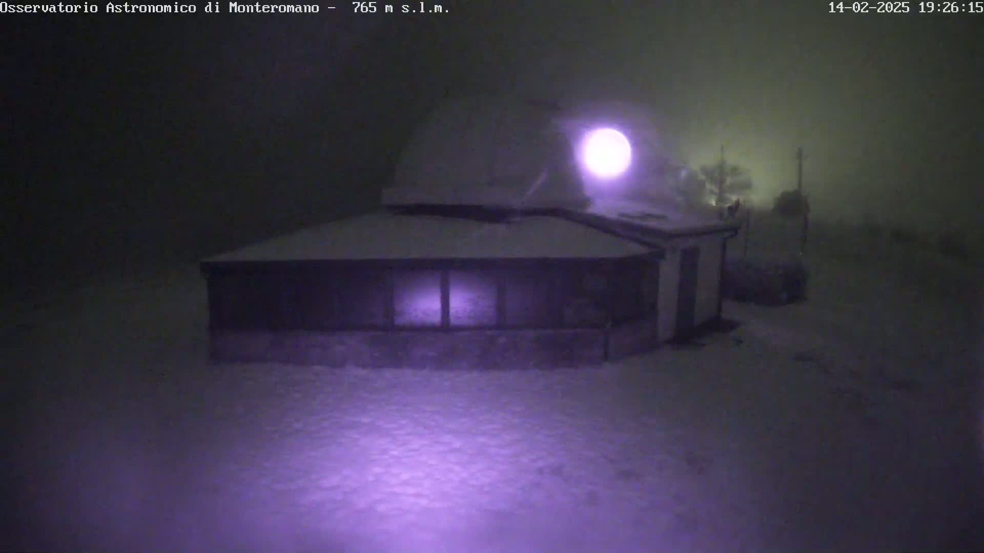 A snow-covered building with a dome on top is illuminated by a light at night in snowy conditions.