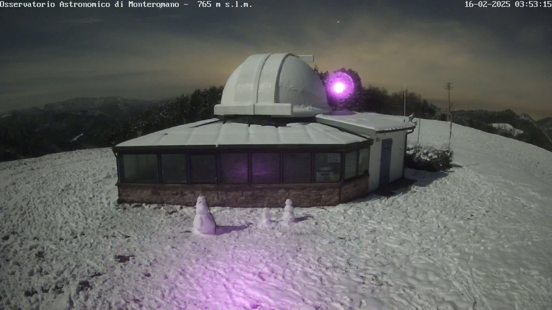 A snow-covered observatory building with a dome sits on a snow-covered hill under a night sky.