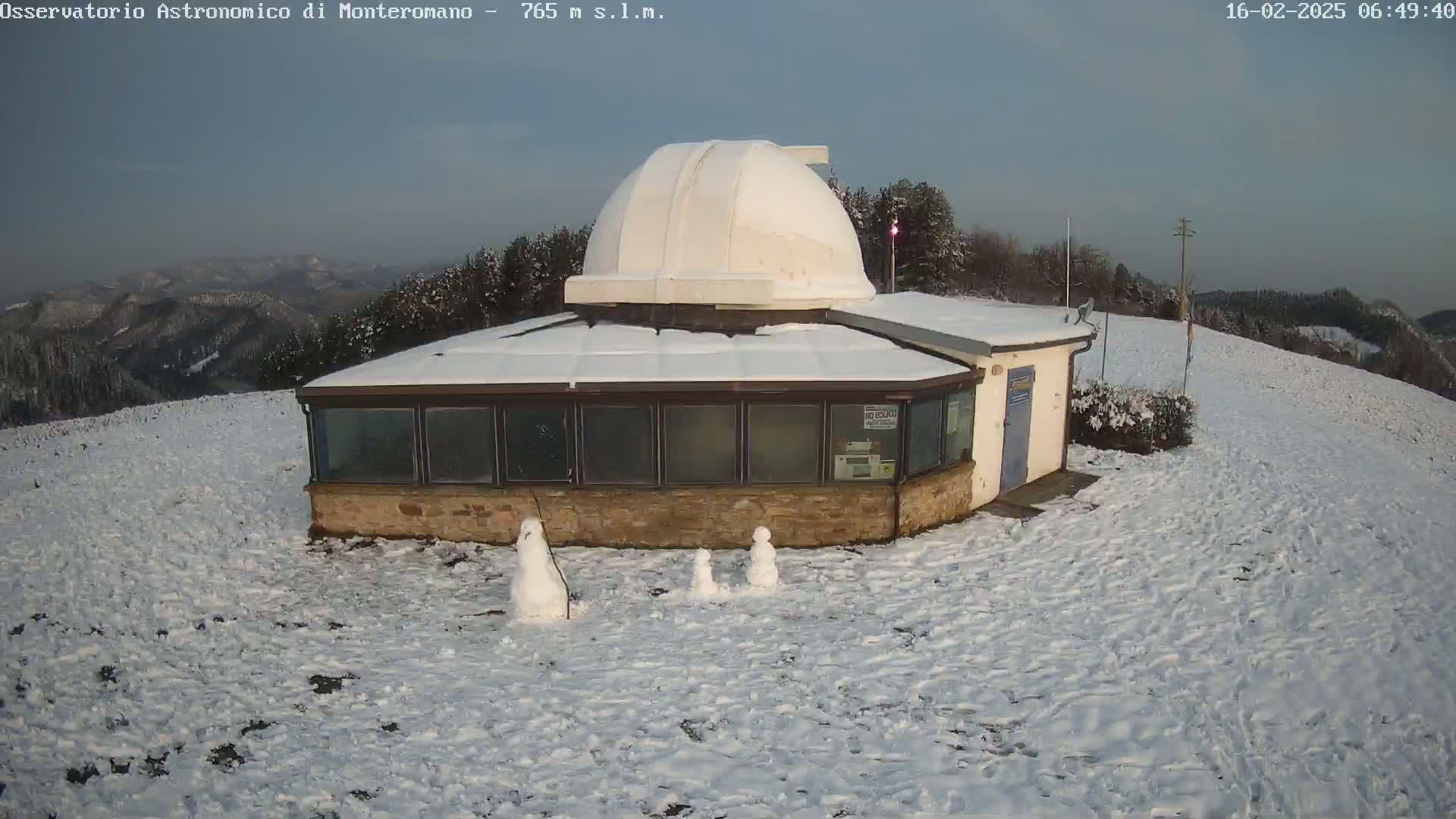 A snow-covered observatory building with a dome sits on a hillside, three small snowmen stand in the snow in front of it, under a clear, light blue sky.