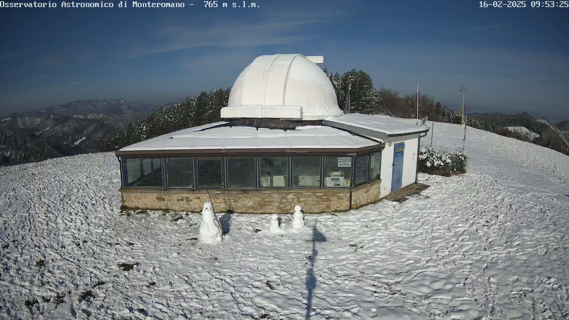 A snow-covered observatory building with a dome sits atop a snow-covered hill, with three small snowmen in front of it under a clear blue sky.