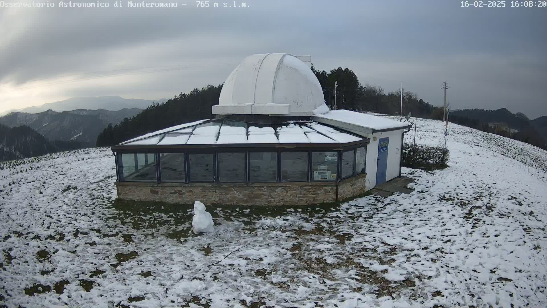 A snow-covered observatory building with a dome sits on a hillside overlooking a mountainous landscape under a cloudy sky.