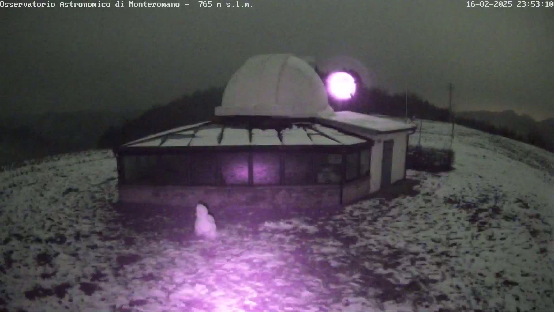 A snow-covered observatory building with a dome is illuminated by a purple light at night under a dark sky.