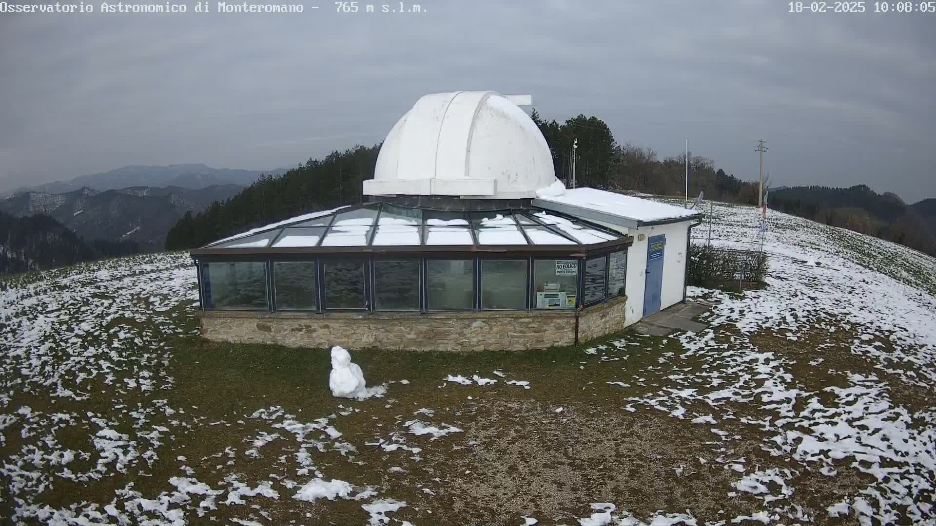 A snow-covered observatory building with a dome sits on a hilltop overlooking a mountain range under an overcast sky.