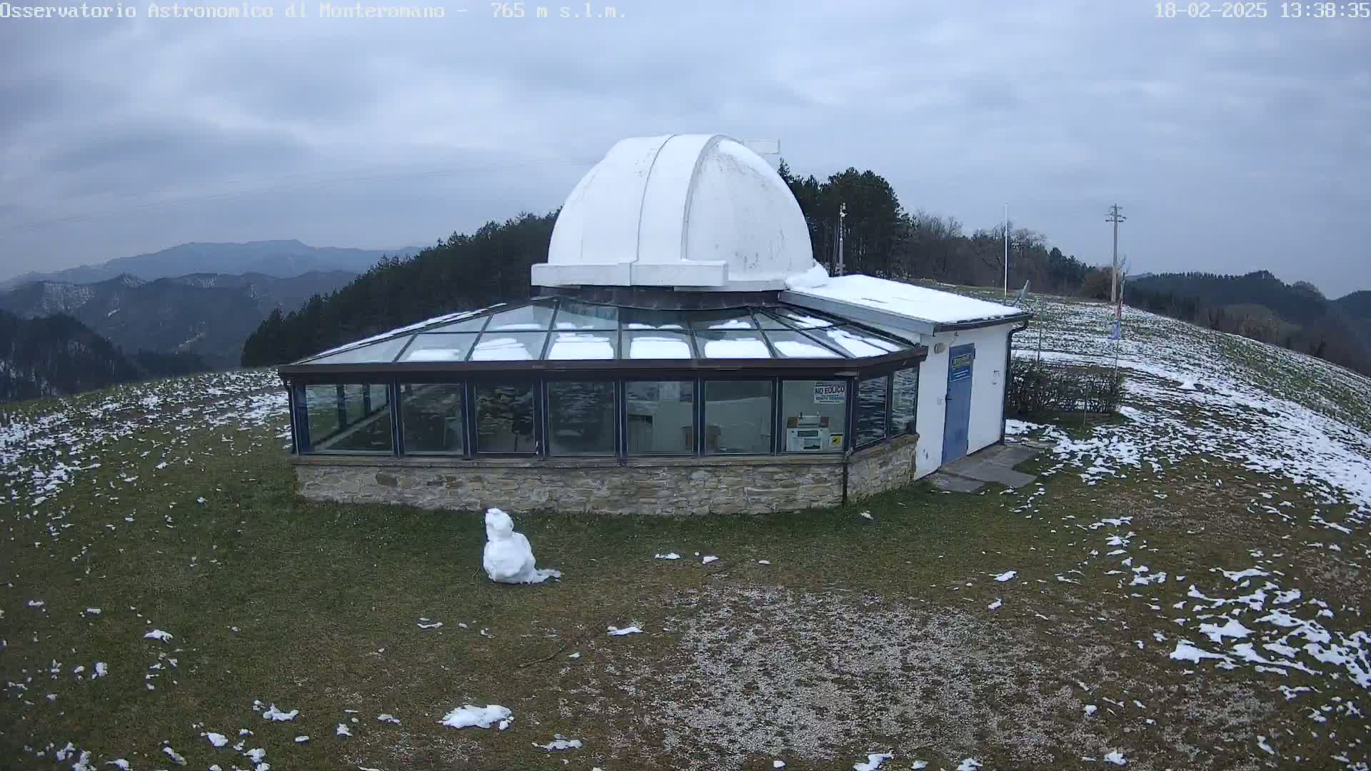 A snow-dusted observatory with a glass sunroom and a dome sits atop a hill overlooking a snow-speckled landscape under an overcast sky.